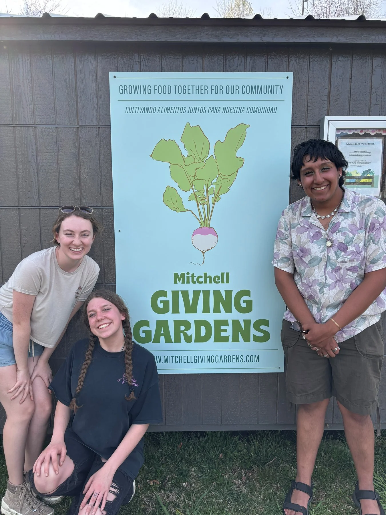 Three people standing in front of a sign for Mitchell Giving Gardens, which features an illustration of a radish with green leaves and a white root. The sign promotes growing food for the community, with text in both English and Spanish.
