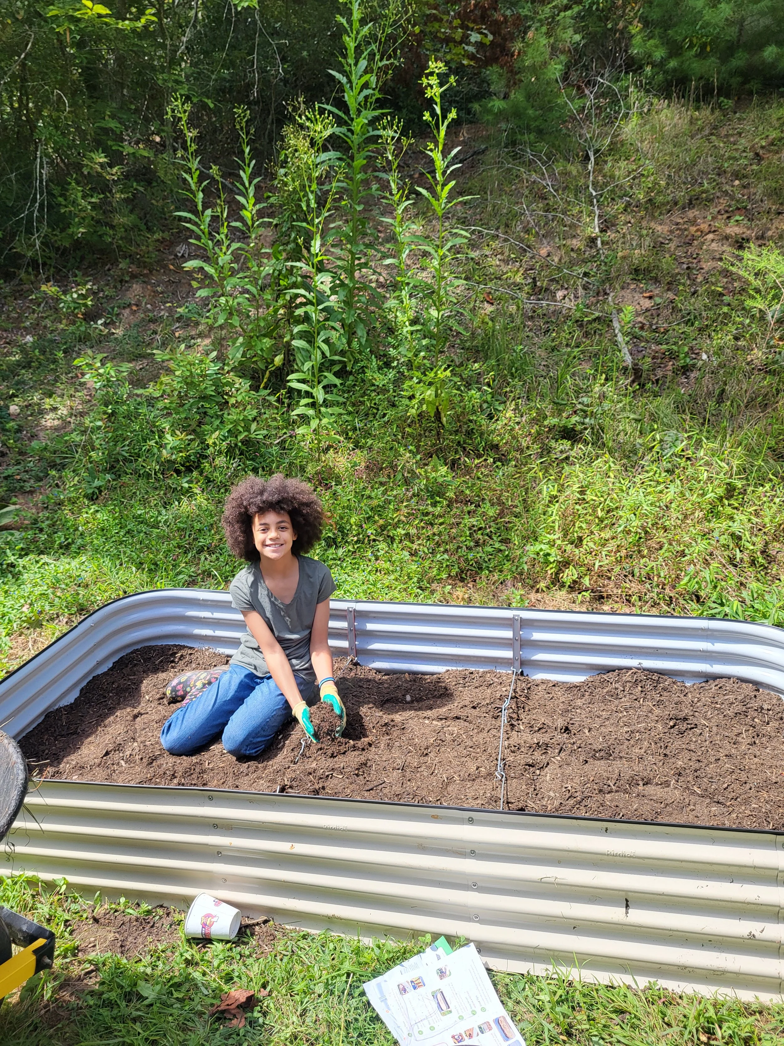 A young girl planting a seed in a garden bed with dirt, surrounded by green plants and trees.