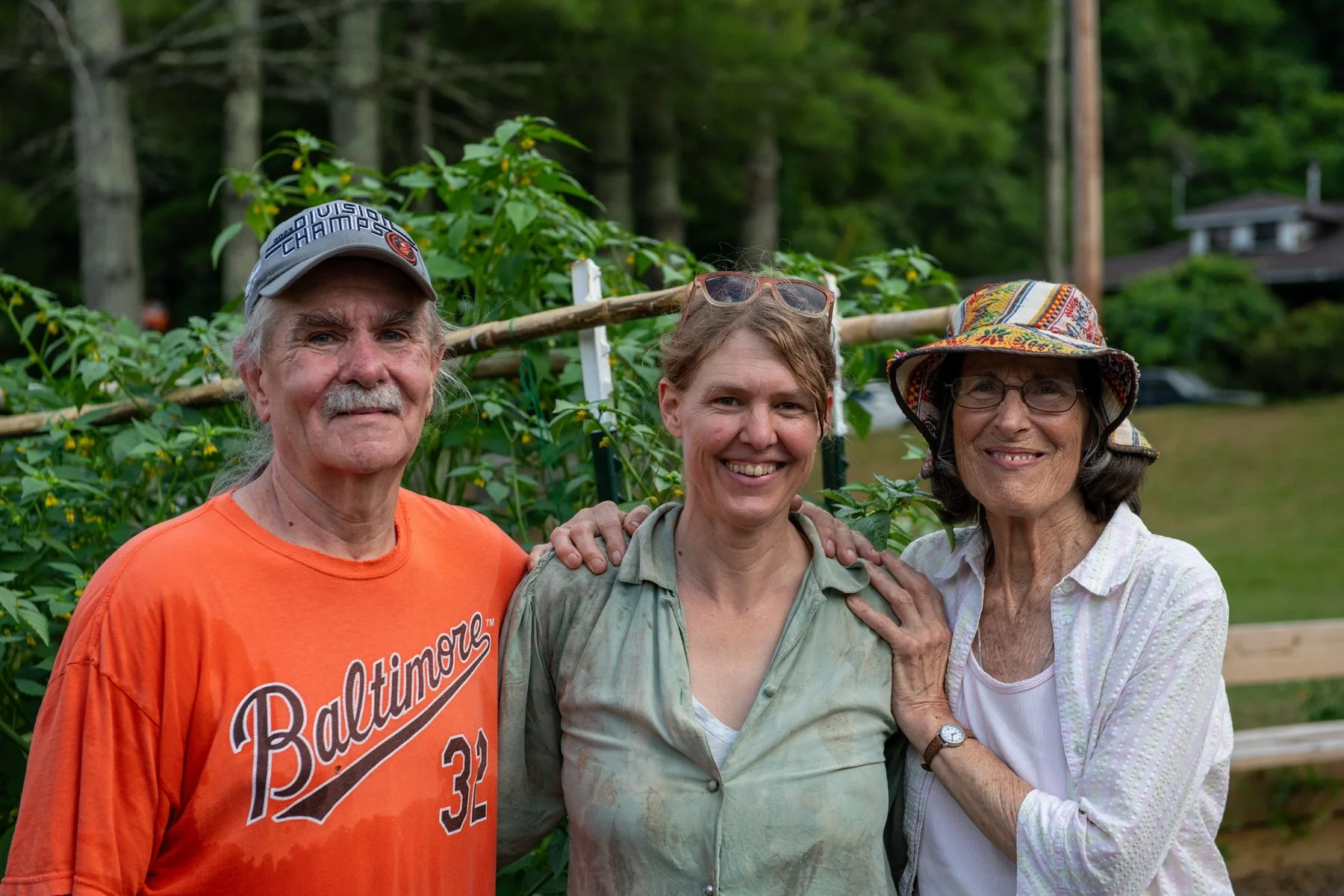 Three smiling people standing outdoors in a garden with greenery and trees in the background; two women and one man, with one woman and the man having their arms around the other woman.