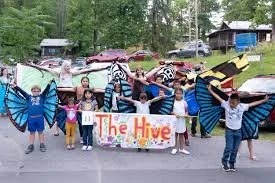 Group of children and adults holding a banner that says 'The Hive,' with some dressed in butterfly costumes, outdoors with trees and parked cars in the background.