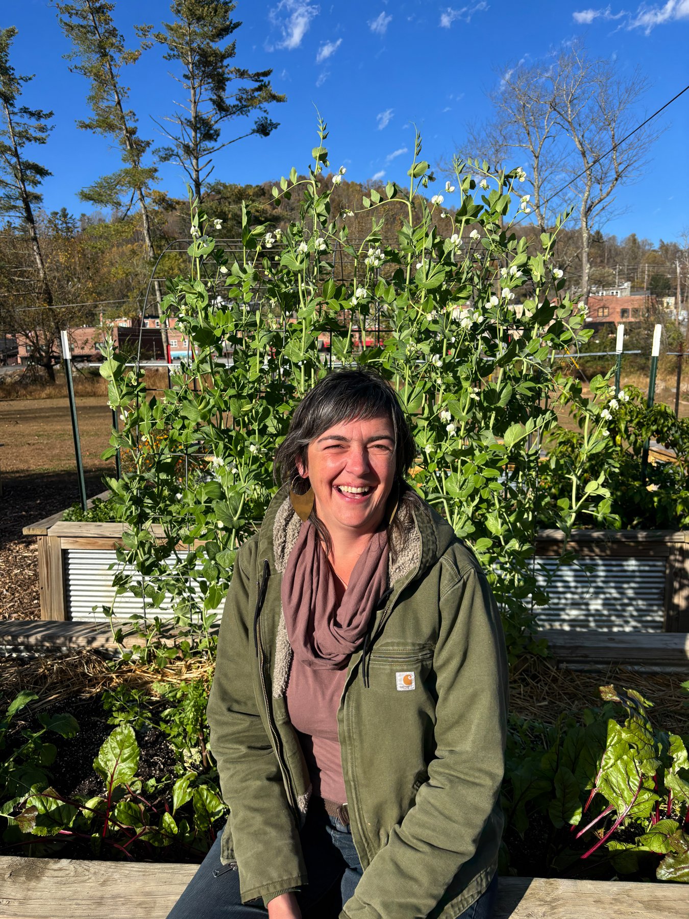 Woman smiling and sitting outdoors in a garden with green plants and trees, blue sky in the background.