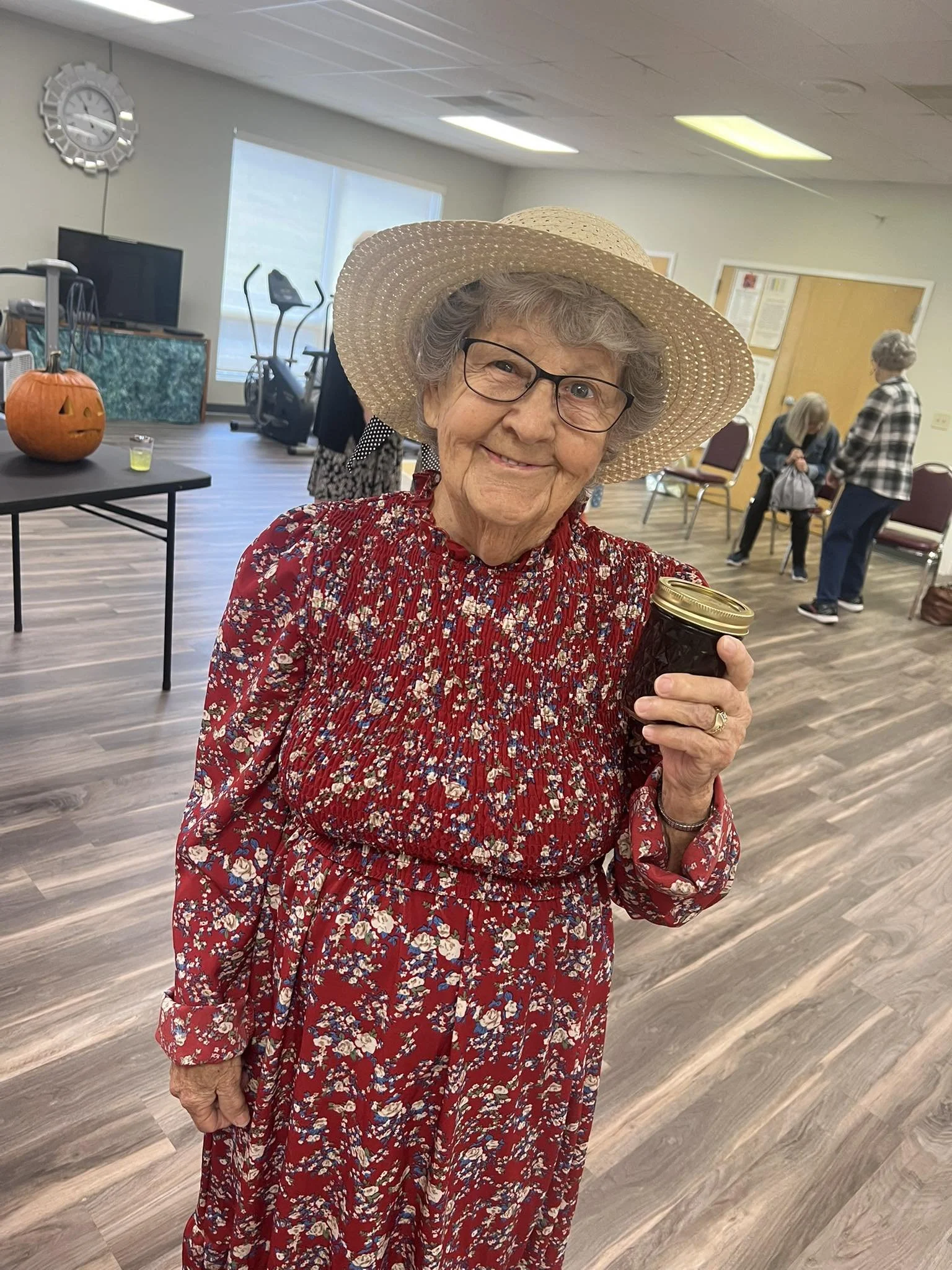 An elderly woman wearing a wide-brimmed straw hat, glasses, and a red floral dress is holding a black jar with a gold lid. She is smiling and indoors at a social gathering.