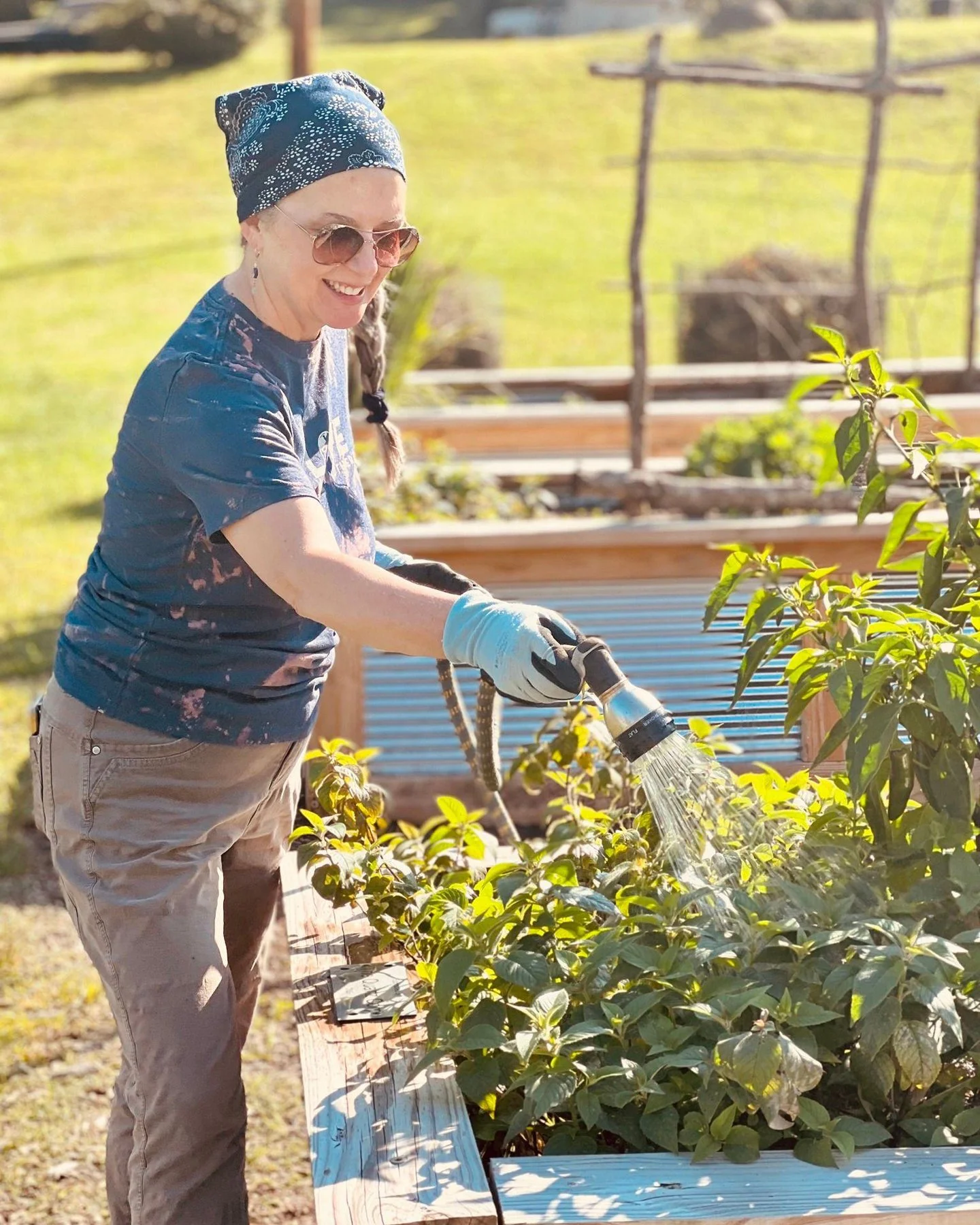 A woman wearing sunglasses, a blue bandana, and gloves watering plants in a the Mitchell Giving Garden beds on a sunny day.