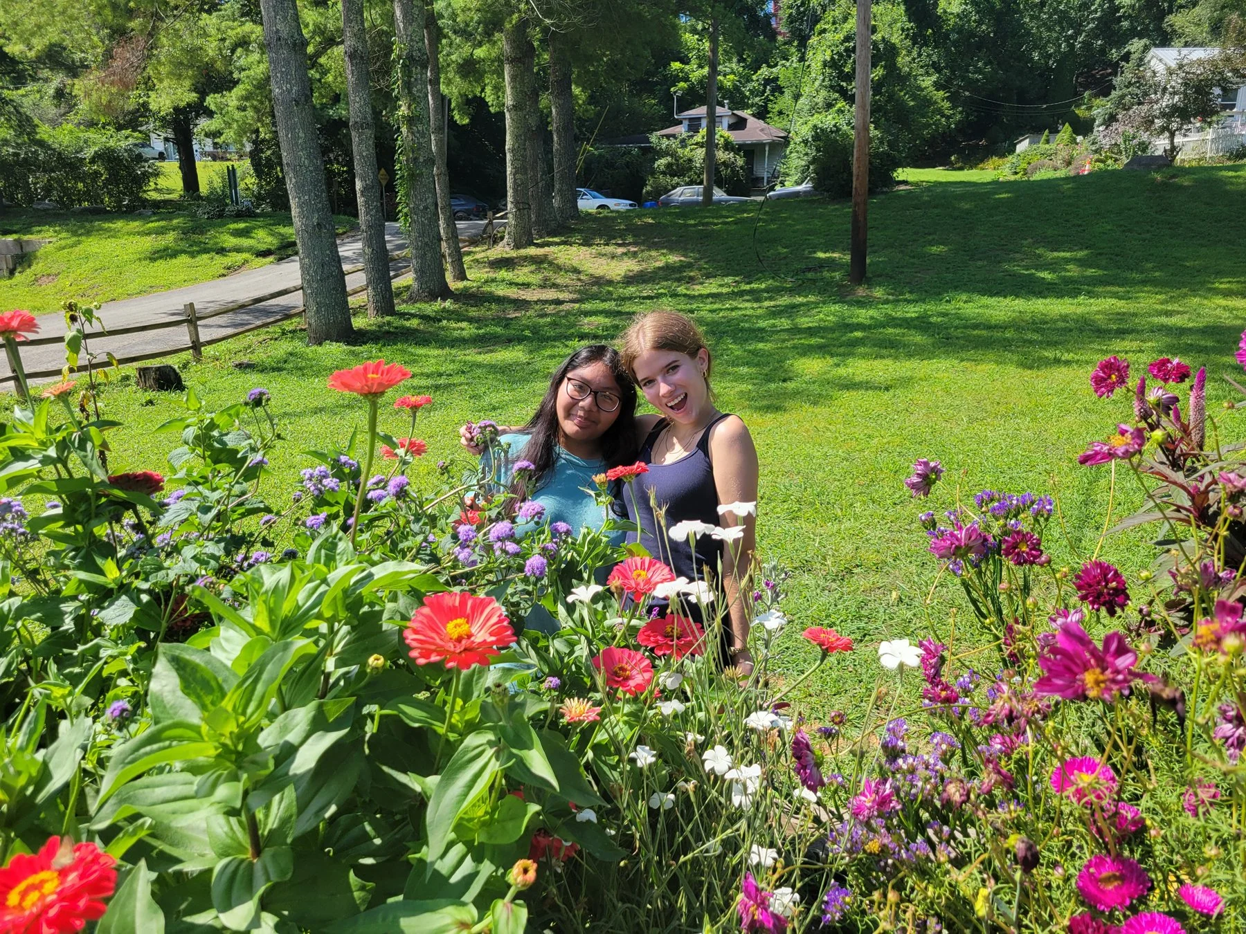 Two girls standing in a garden with colorful flowers, trees, and a grassy area.