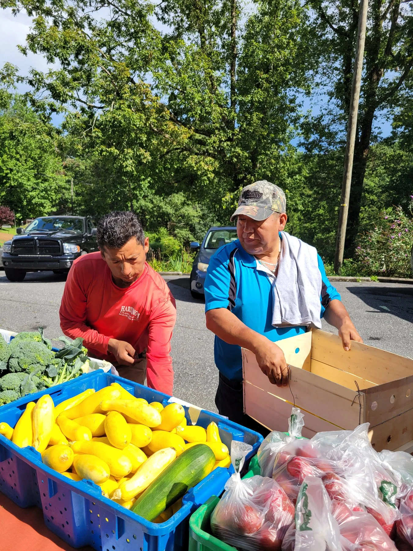Two men at an outdoor farmers market stand with fresh vegetables including yellow squash, zucchini, broccoli, and tomatoes.