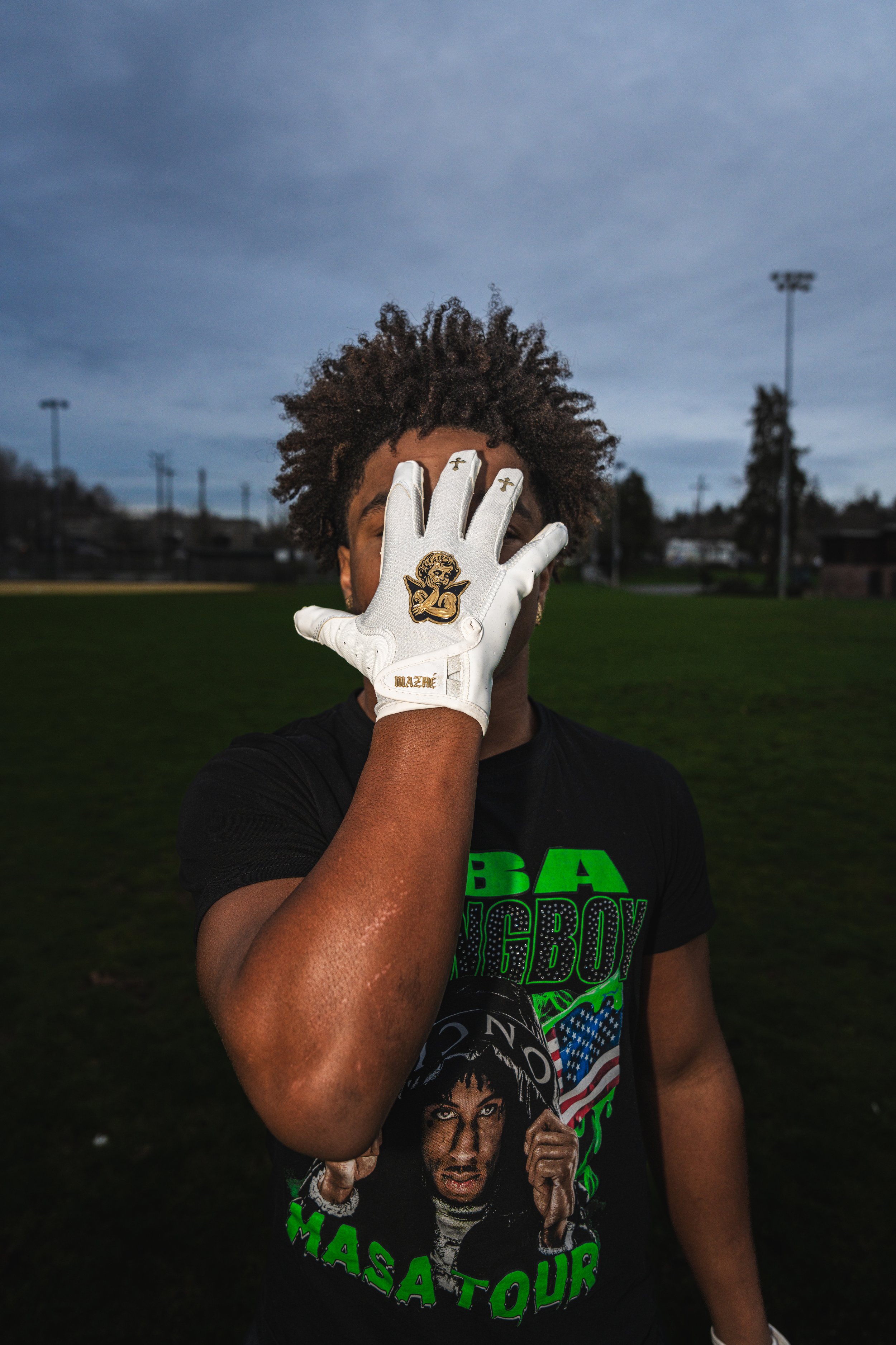 A person with curly hair wearing a black T-shirt with a graphic and text, holding up their right hand covered in a white glove with a cartoon character patch, standing outdoors on a field under a cloudy sky.