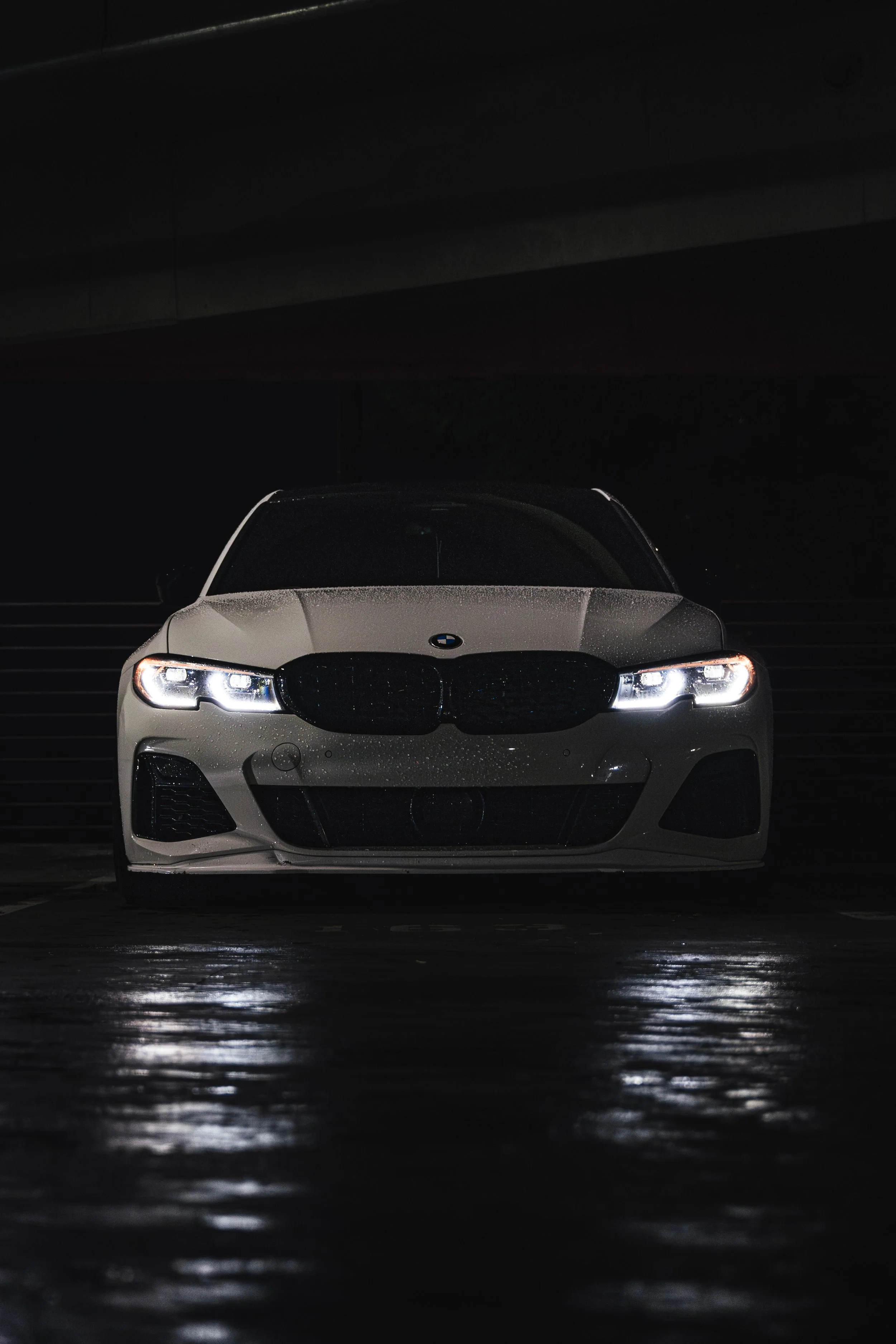 Front view of a white BMW sedan with headlights on, parked in a dark, wet environment.