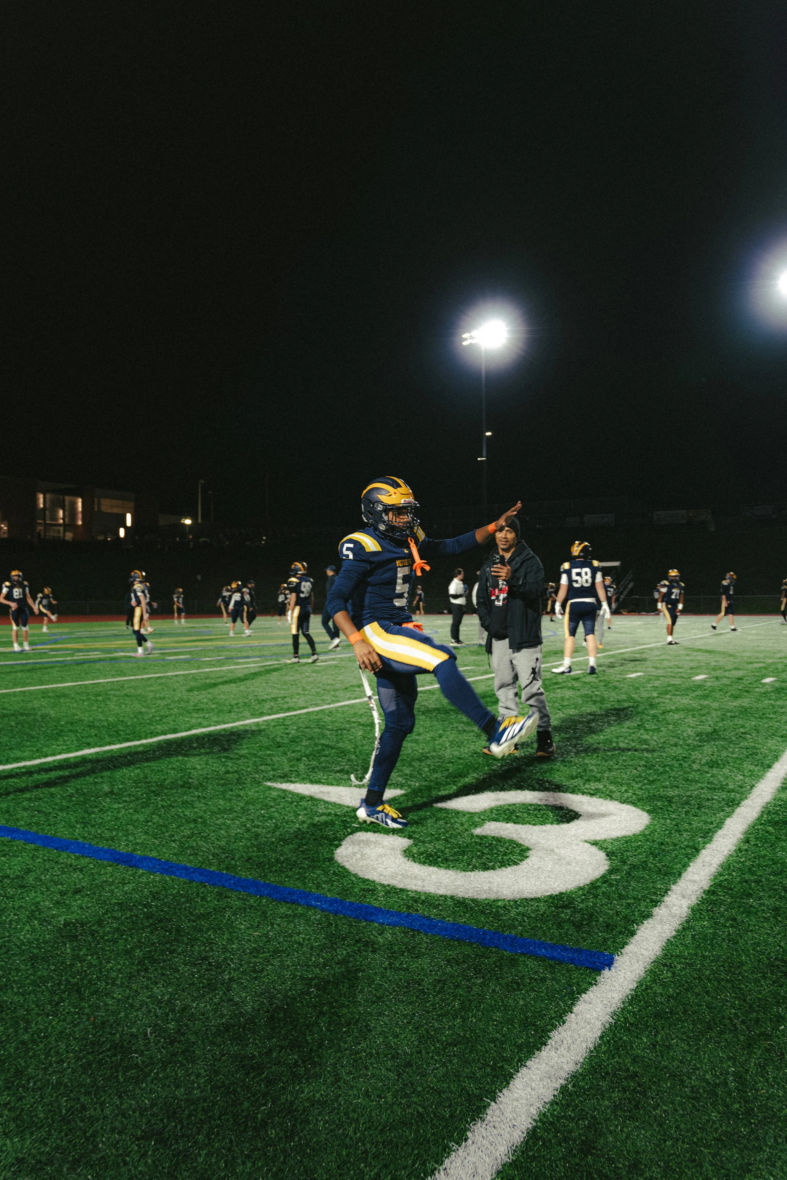 A football player wearing a dark blue and yellow uniform with the number 5 is warming up or stretching on the field under floodlights at night, with other players and a person standing nearby in the background.