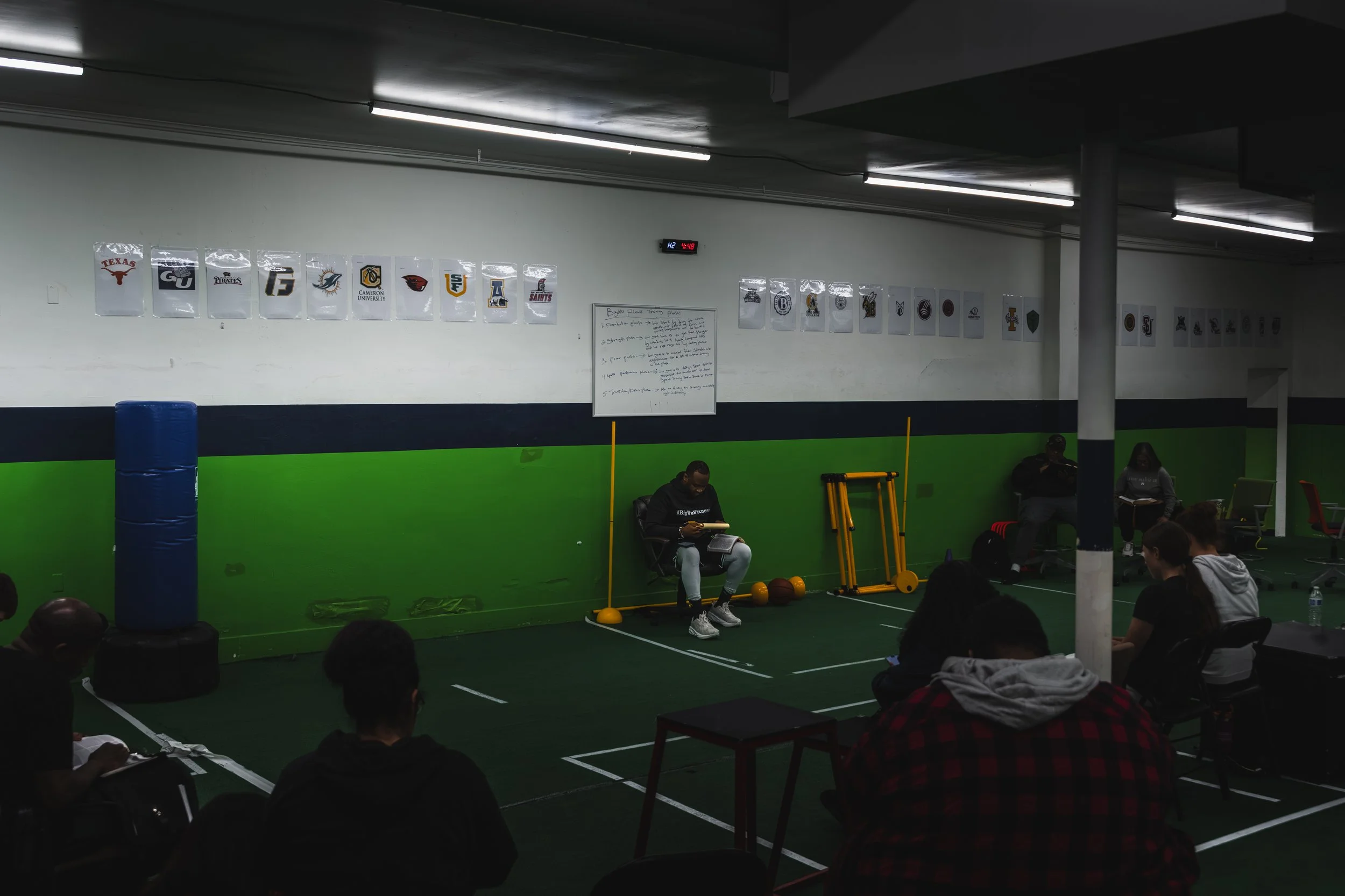Indoor gym with green and white walls, sports logos hanging on the wall, people seated, and a person sitting on a chair with fitness equipment around.