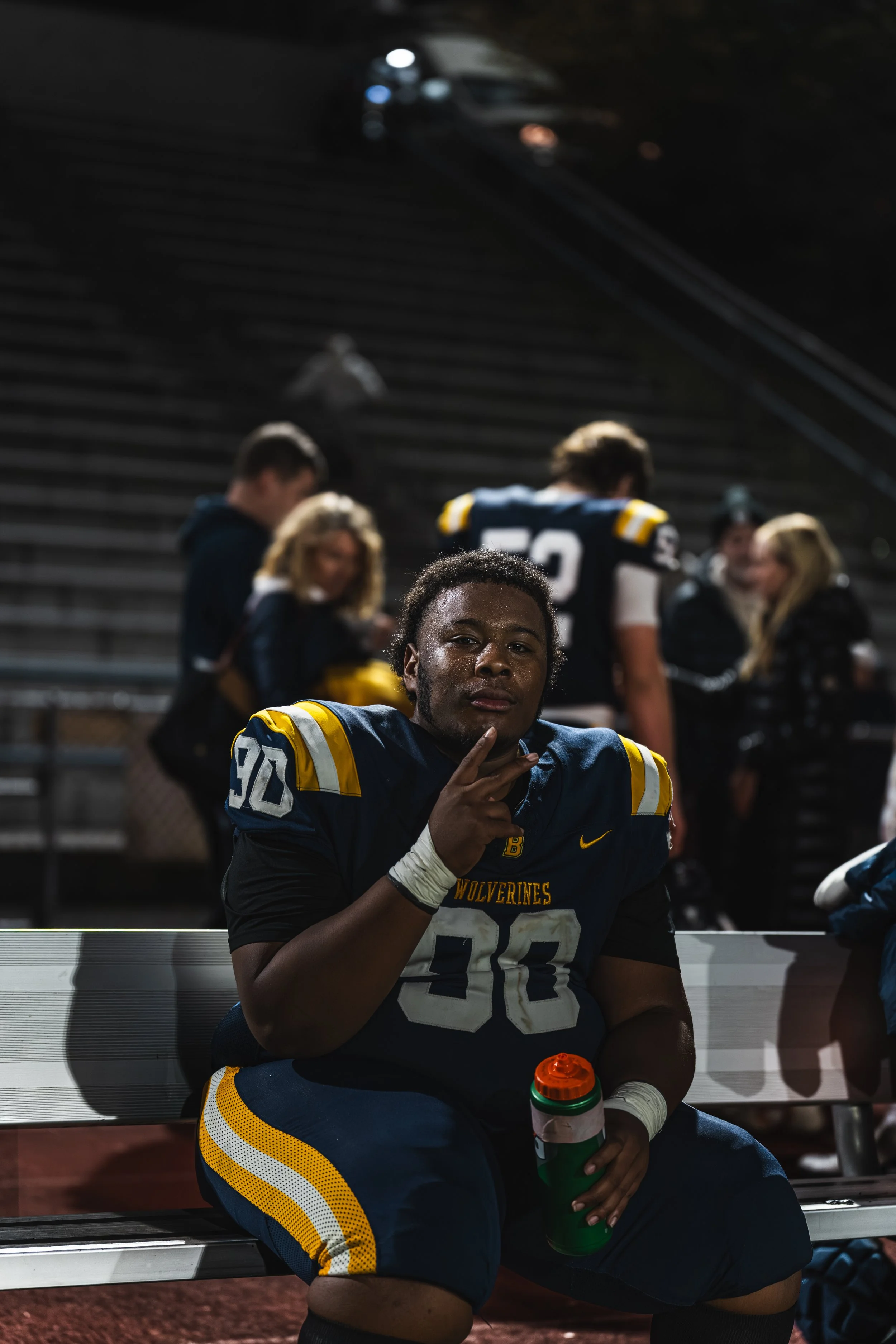 A football player in a blue and yellow uniform sitting on a bench, holding a water bottle, making a peace sign, and looking at the camera with a serious expression.