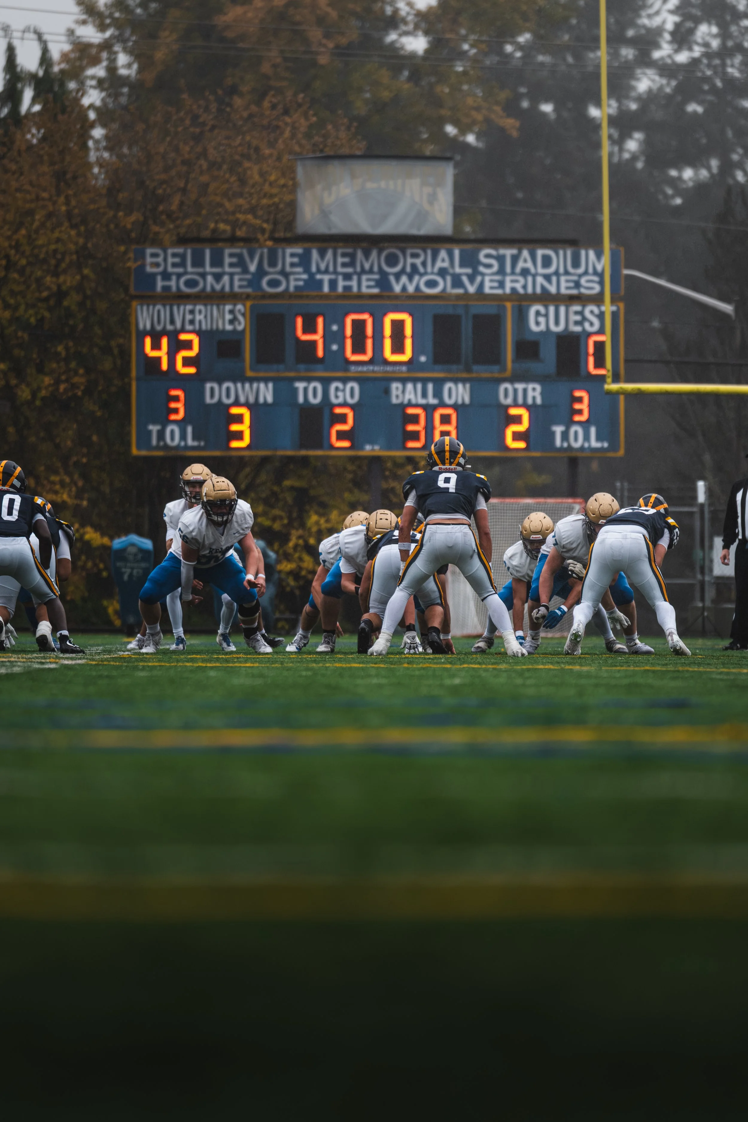 Young football players on the field during a game at Bellevue Memorial Stadium, with a scoreboard in the background displaying 42 to 3 in favor of the Wolverines, four minutes remaining in the game.
