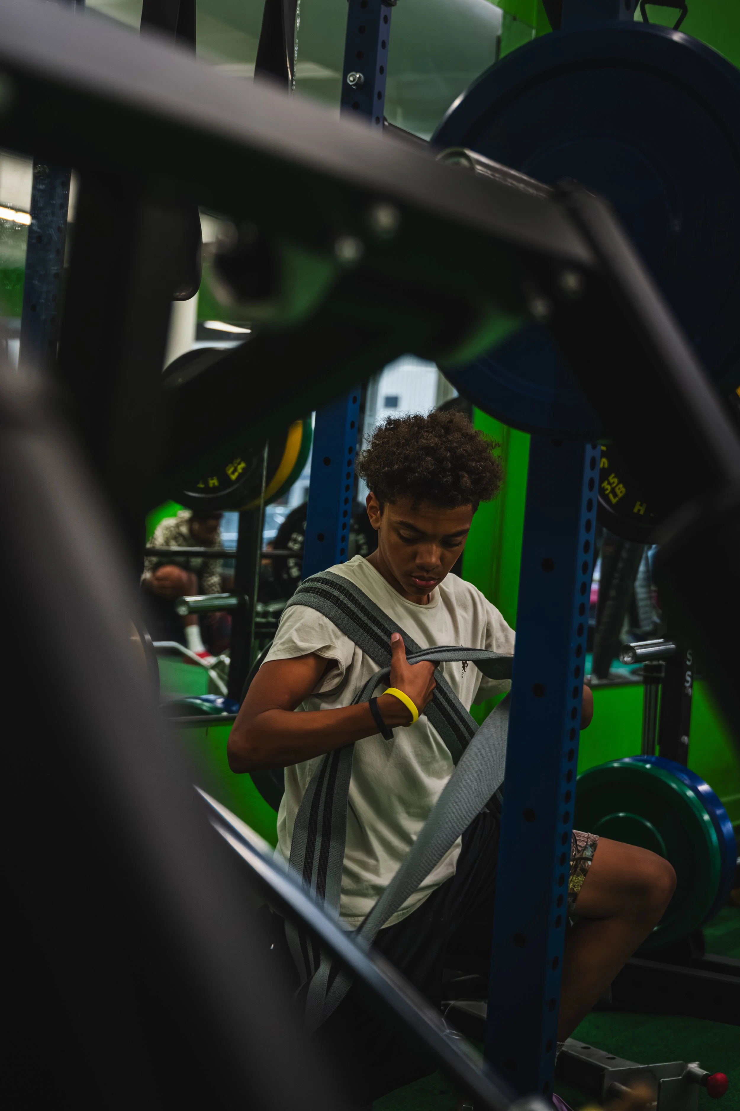 A young man adjusting a weightlifting strap in a gym, surrounded by black weights and green walls.