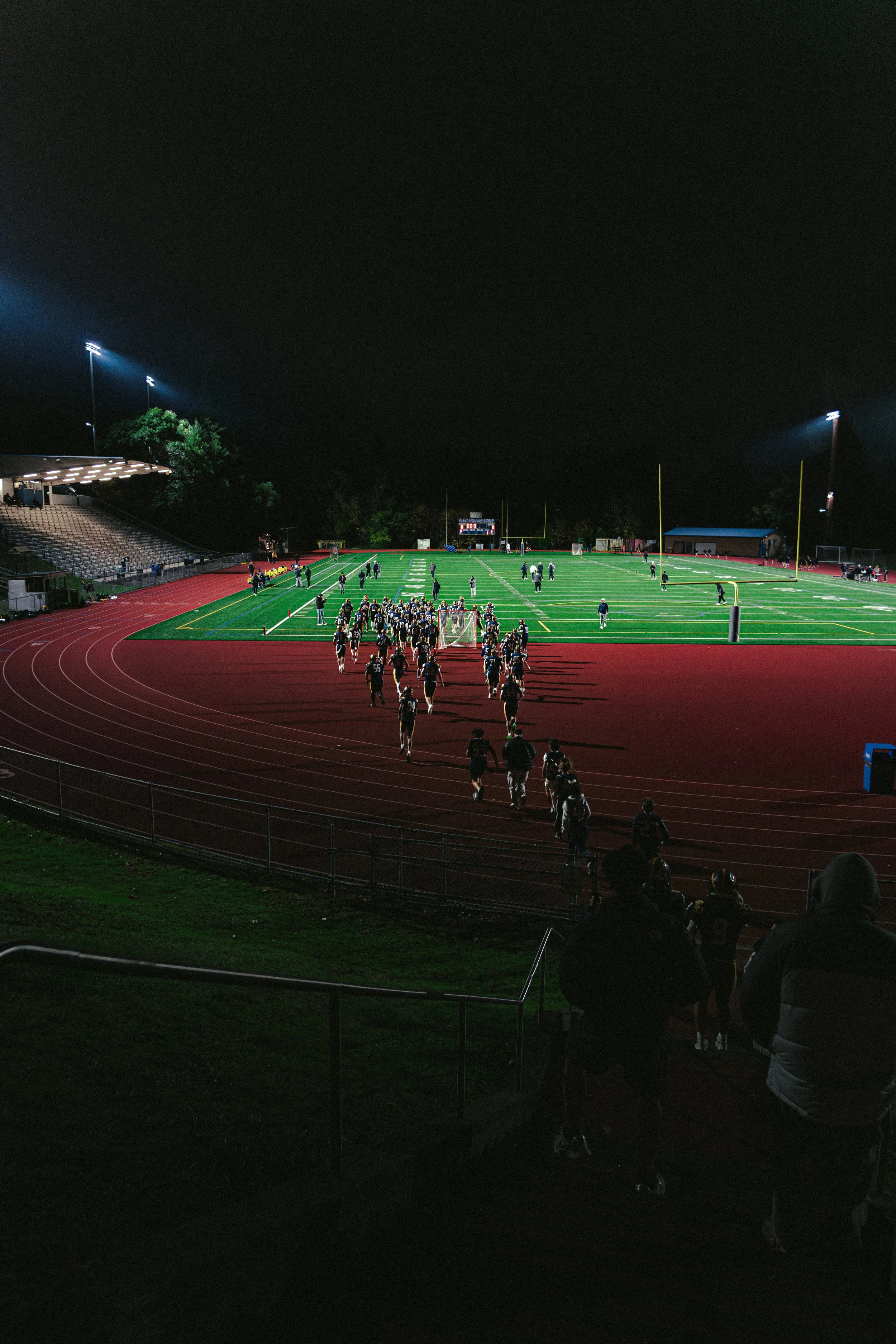 Night football game at a stadium with players on the field and spectators watching from the stands and sidelines.