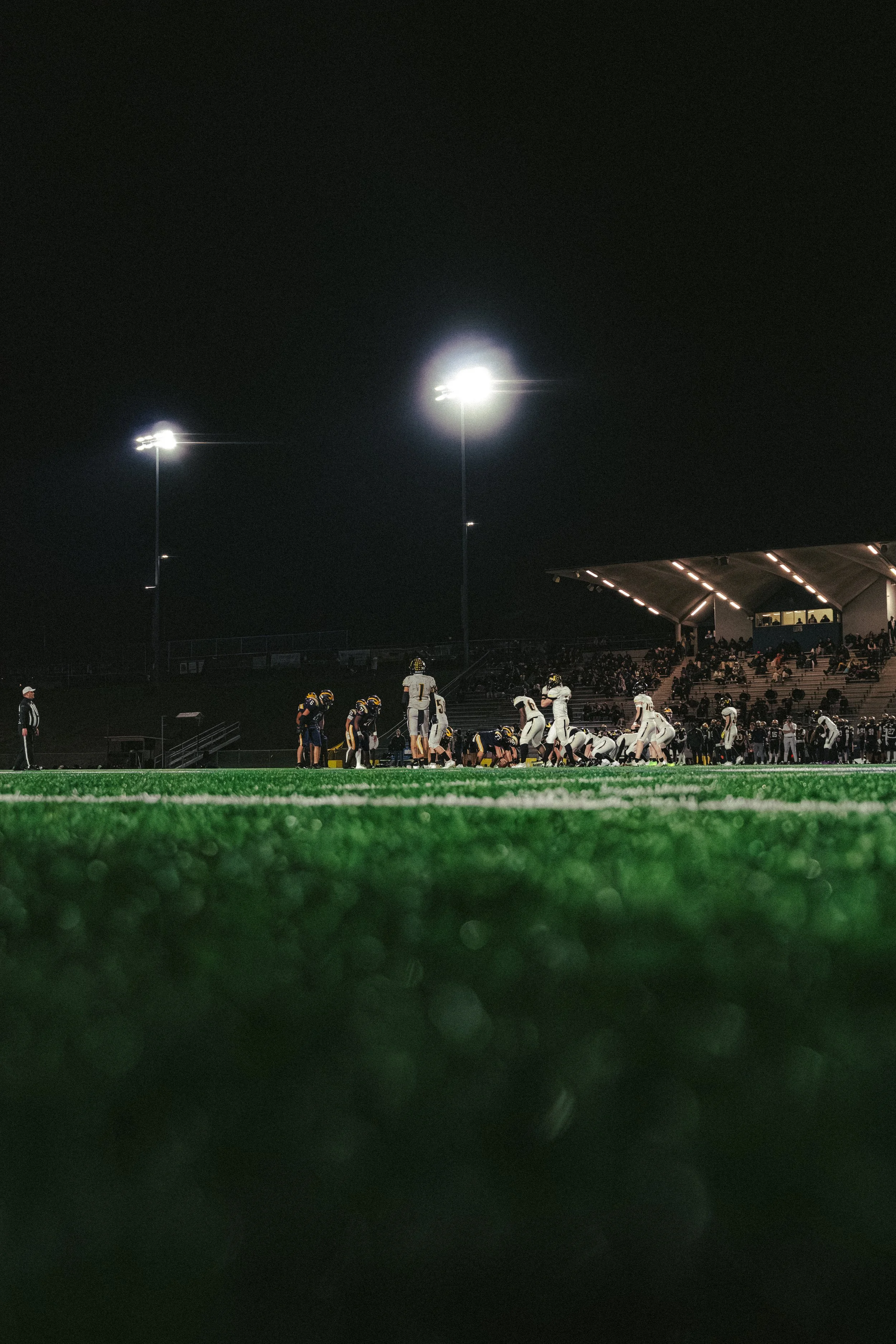 Nighttime football game under stadium lights with players on the field and spectators in the stands.