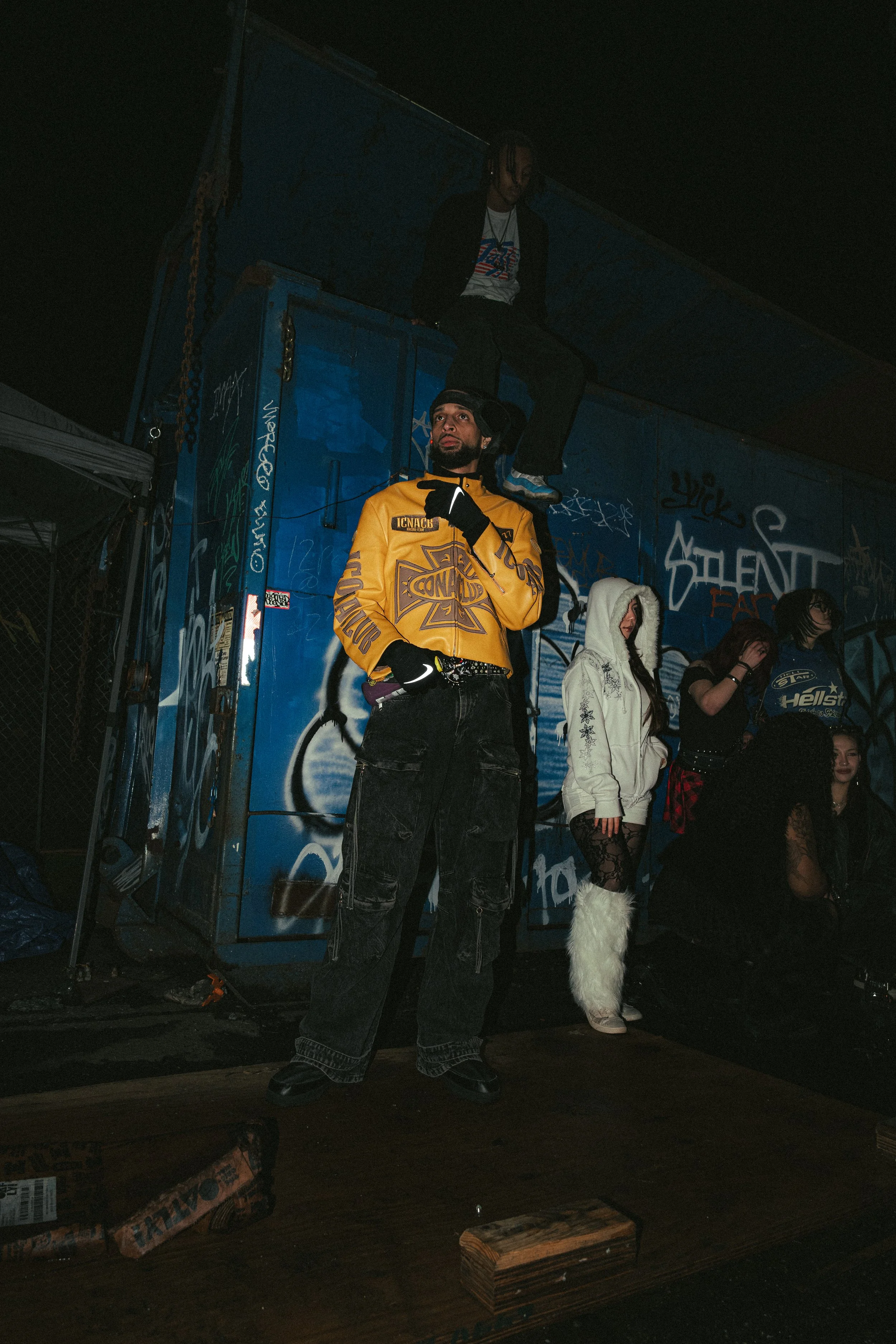 A group of young people at an outdoor night event, with graffiti-covered blue storage containers in the background. One person stands in front, wearing a yellow jacket and black gloves, while another sits on top of the container. Others are standing 