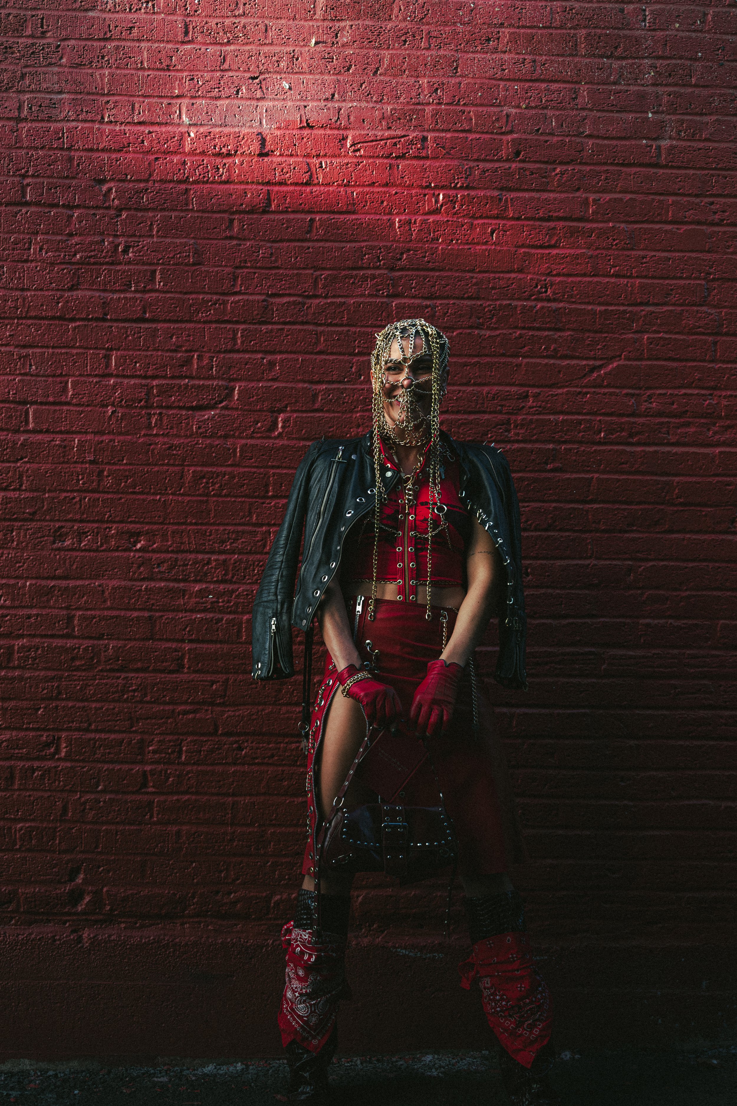 A person standing against a red brick wall, wearing a leather jacket, red outfit with chain accessories, face mask with chains, and bandana on their legs.