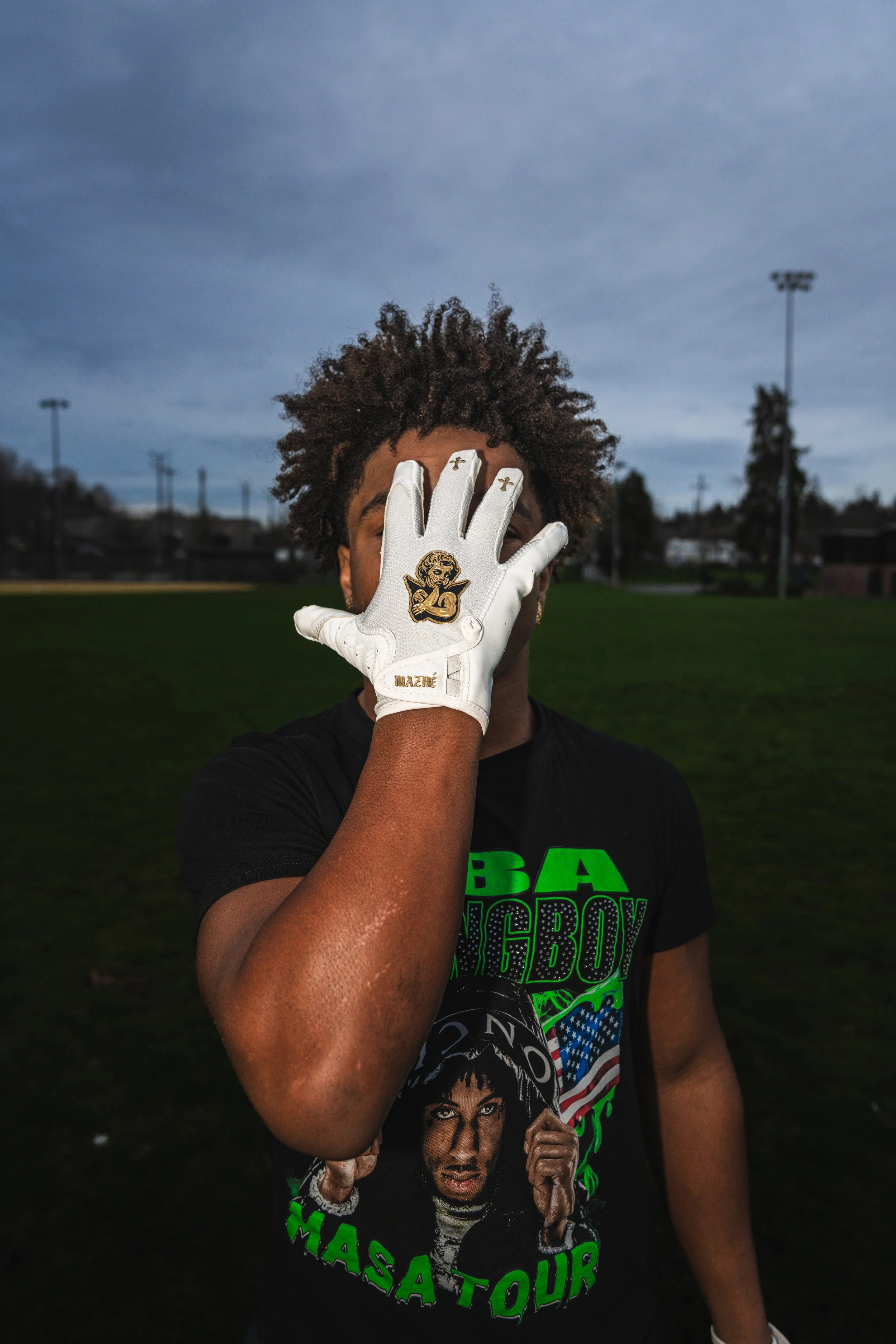 Person standing outdoors on a field, partially covering their face with their left hand which is wearing a white glove with a cartoon character patch.