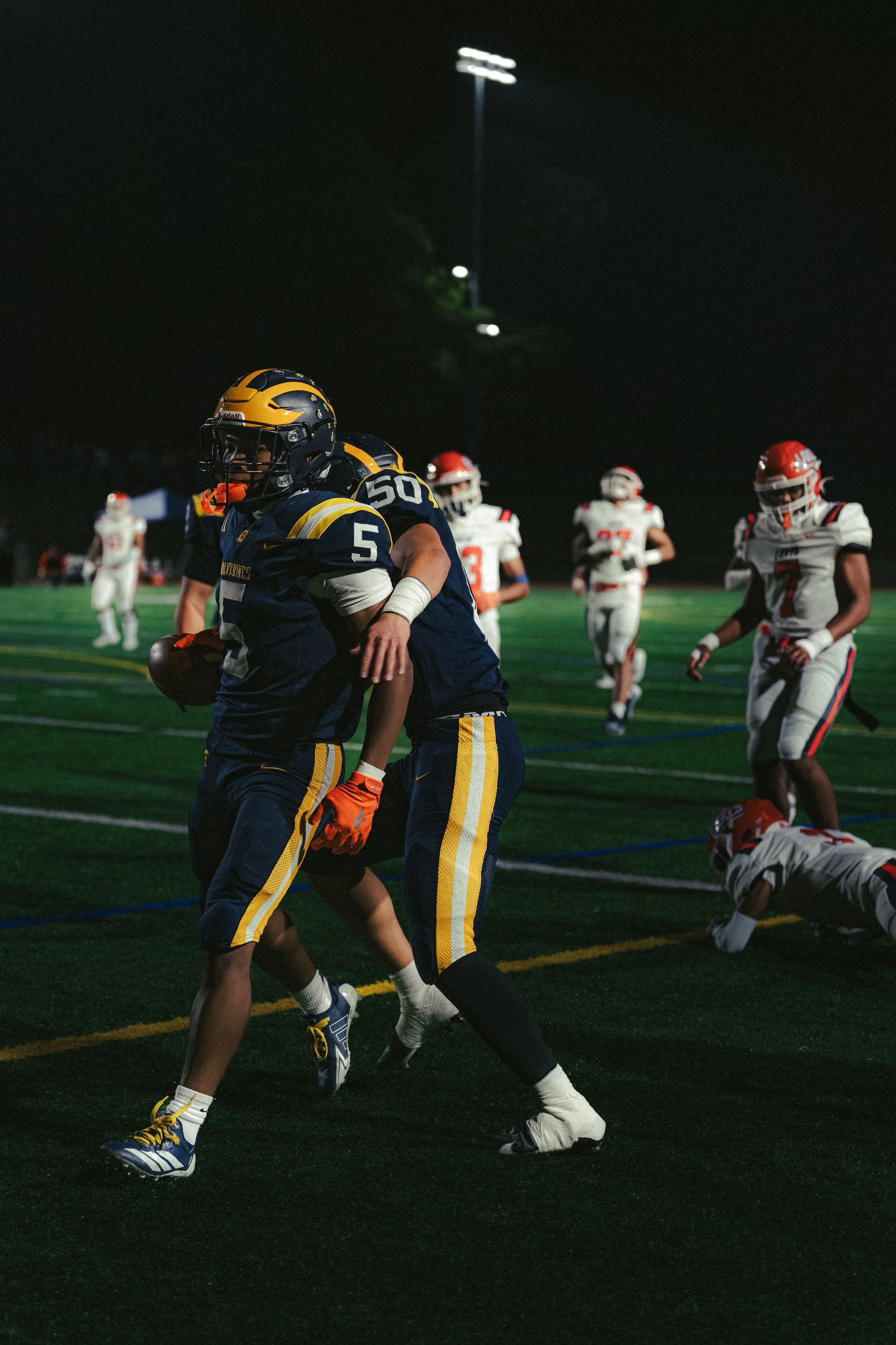 A nighttime football game with players in action on the field. Two players in navy and yellow uniforms are celebrating, while players in white and red uniforms are in the background.