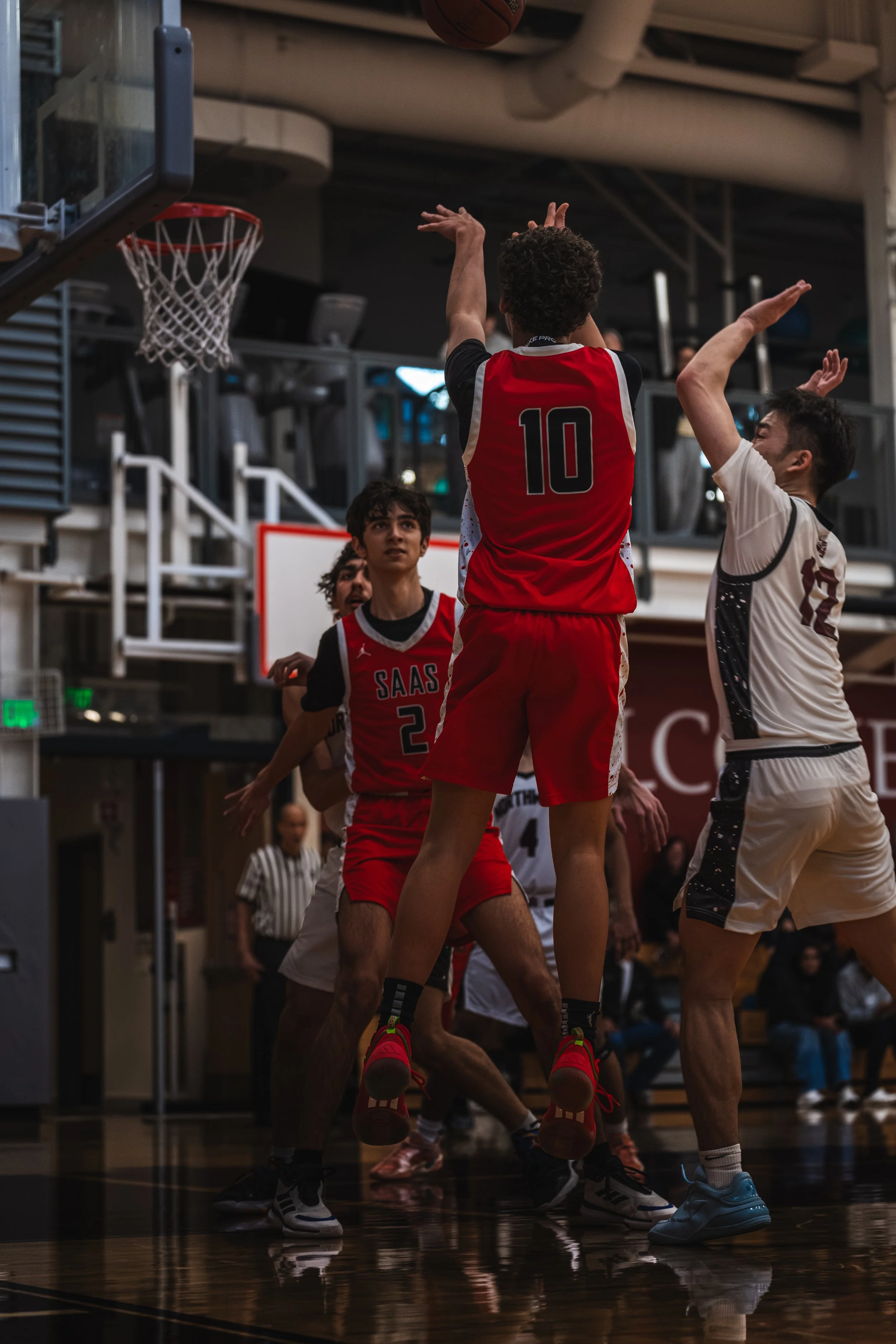 Young basketball players in red and white uniforms engaged in an indoor game, with one player jumping to make a shot near the basket.