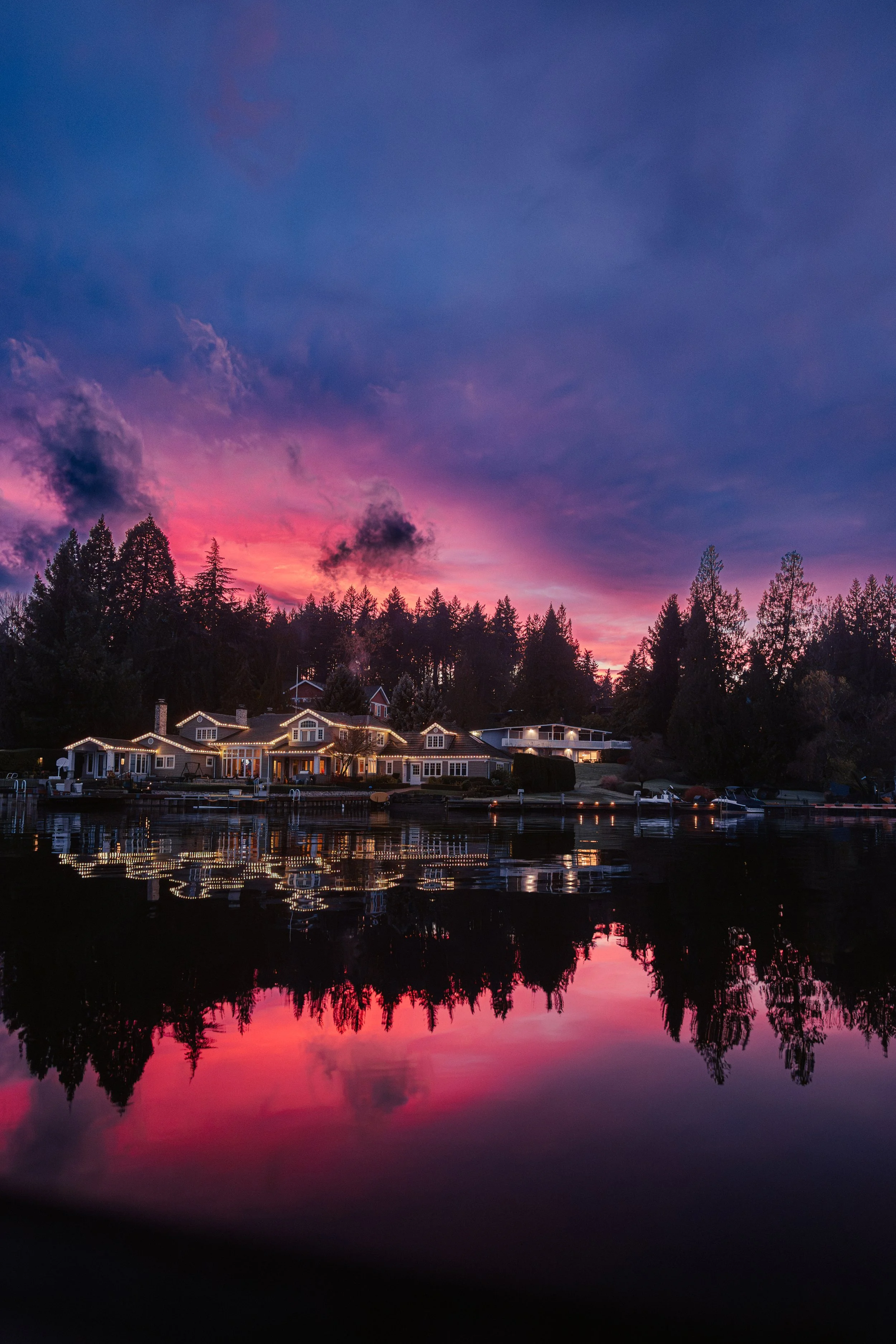 A lakeside scene at sunset with houses decorated with lights, calm water reflecting the colorful sky and silhouetted trees.