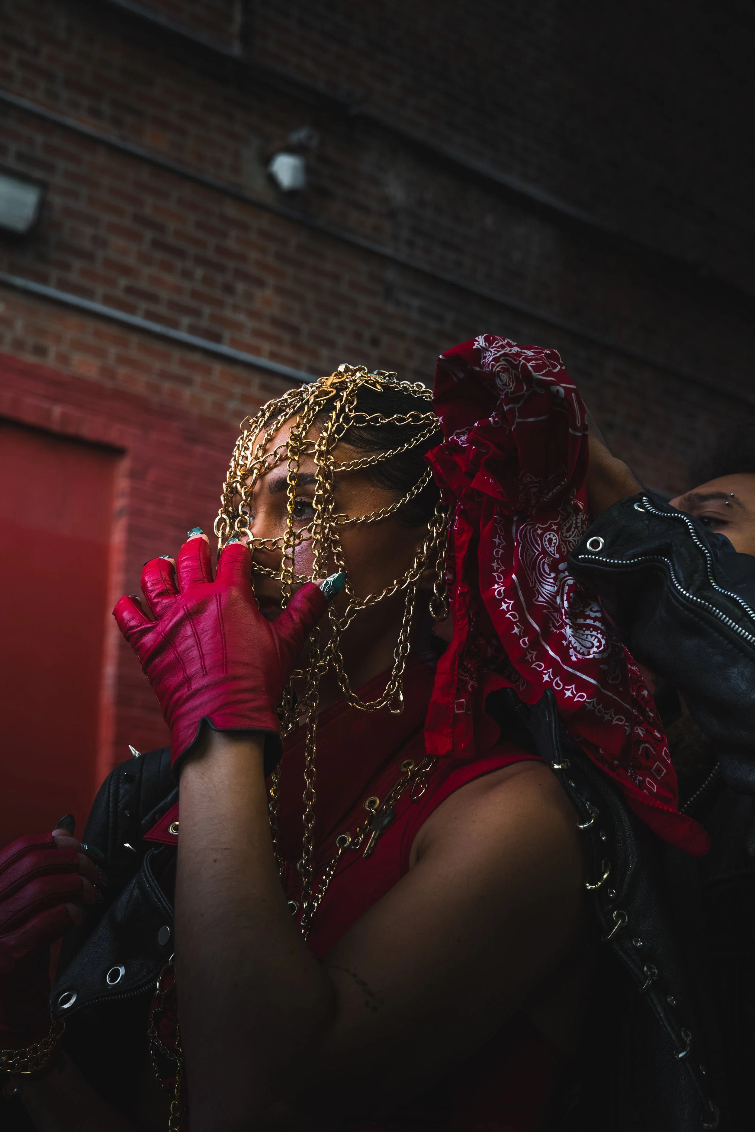 Person dressed in red with gold chains, red bandana, red gloves, and leather jacket, posing outdoors against a brick wall.