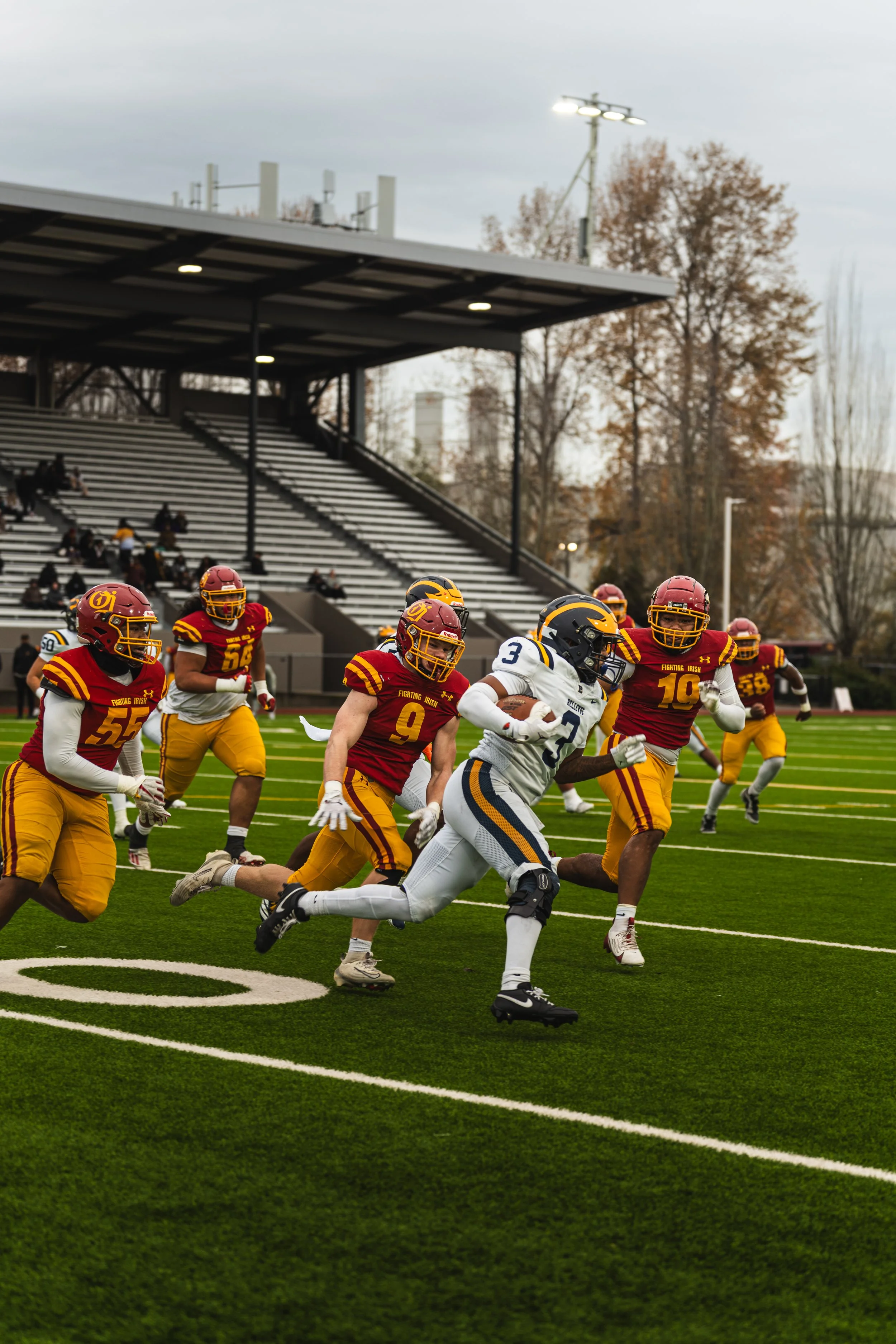 A football game with players from opposing teams on the field, some in white uniforms and others in red and yellow uniforms, with a stadium and empty bleachers in the background.