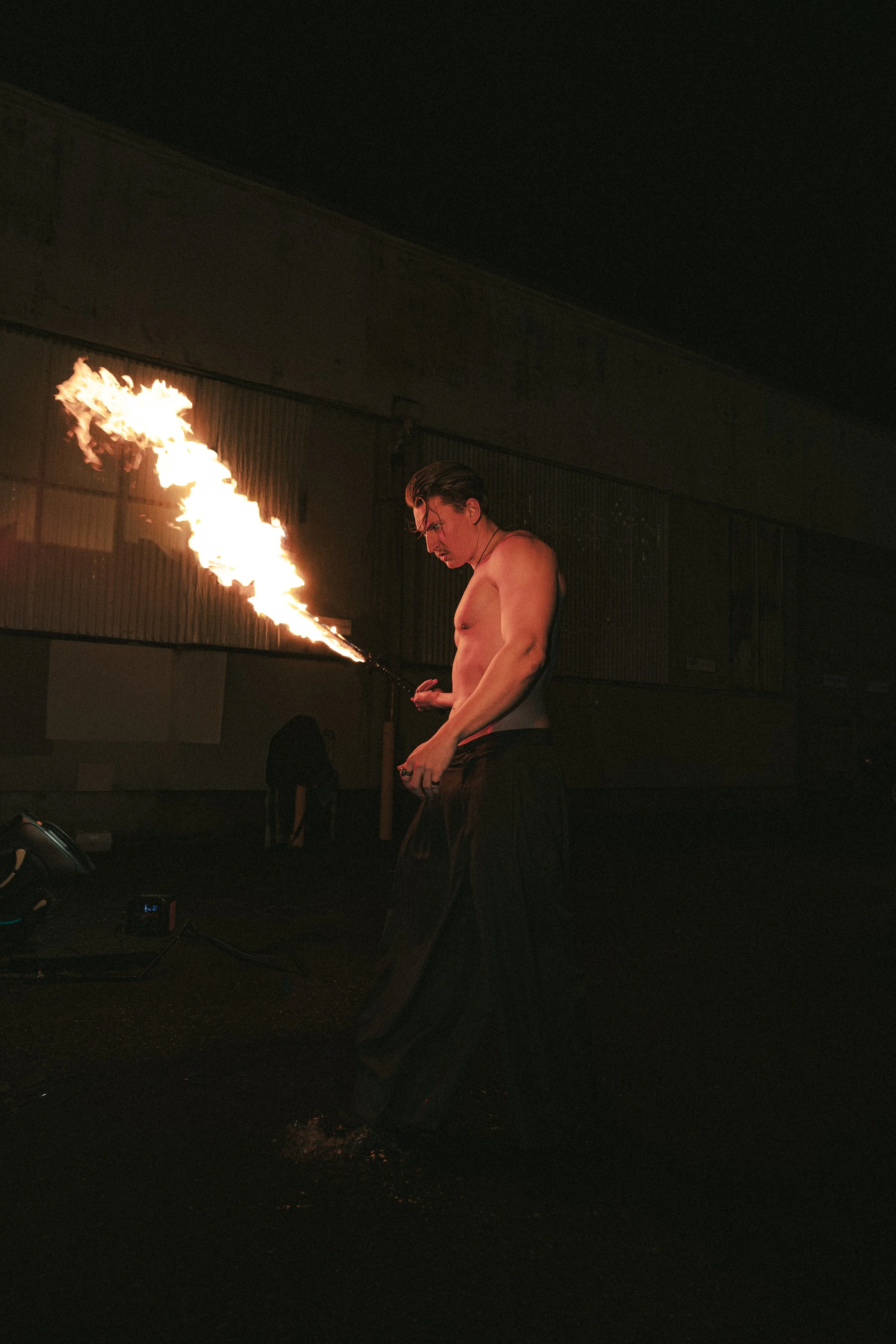 A shirtless male performer holding a fire torch, with a flame in front of a dark industrial background.
