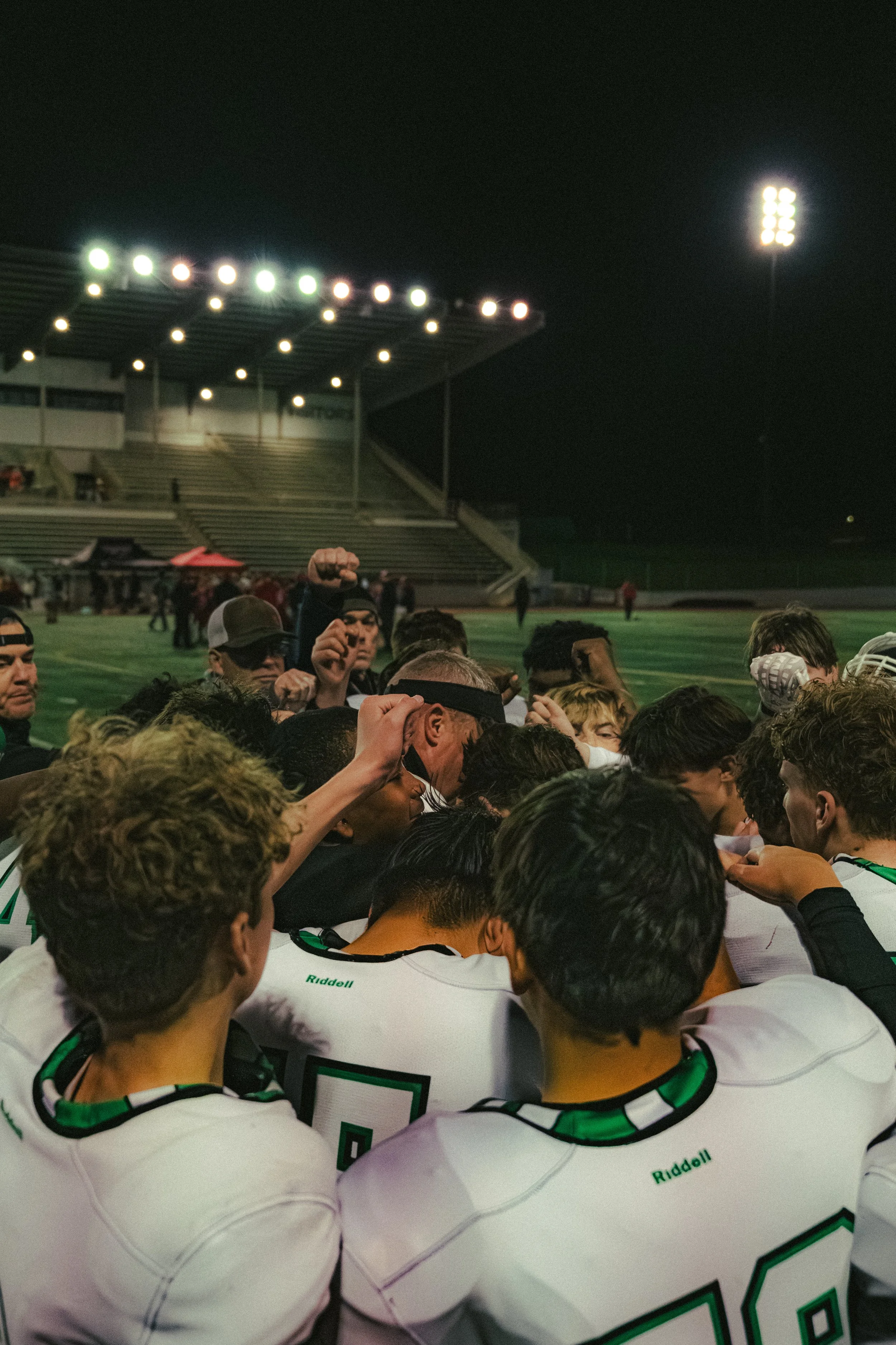 A football team huddles together on the sidelines of a stadium field at night, illuminated by bright stadium lights, with some players raising fists.