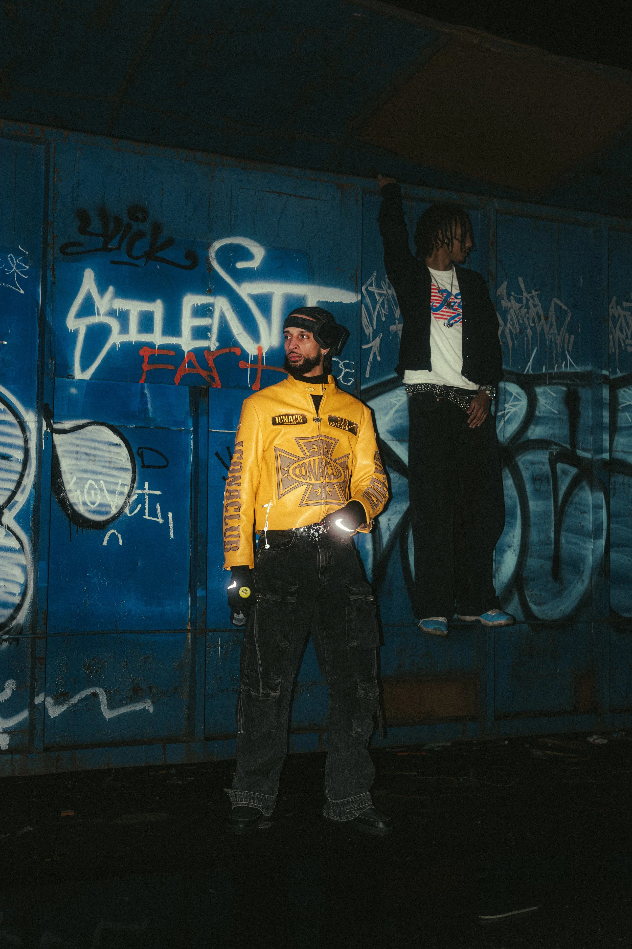 Two young men pose in front of a graffiti-covered wall, one standing with hands in pockets wearing a yellow jacket, and the other hanging from above wearing a black jacket and dark pants.