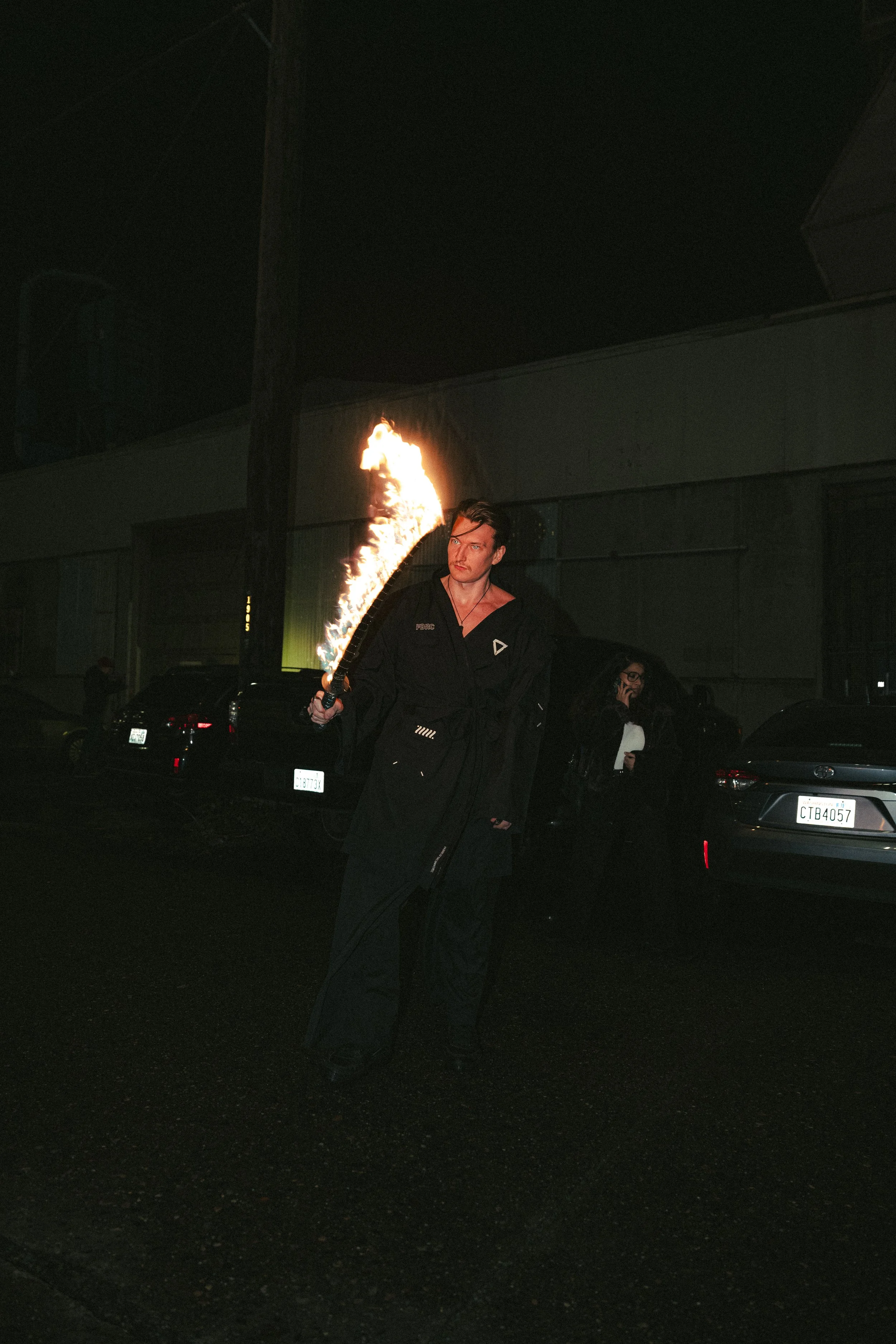 A man holding a flaming torch standing on a dark street at night, with cars parked in the background and a woman talking on her phone.