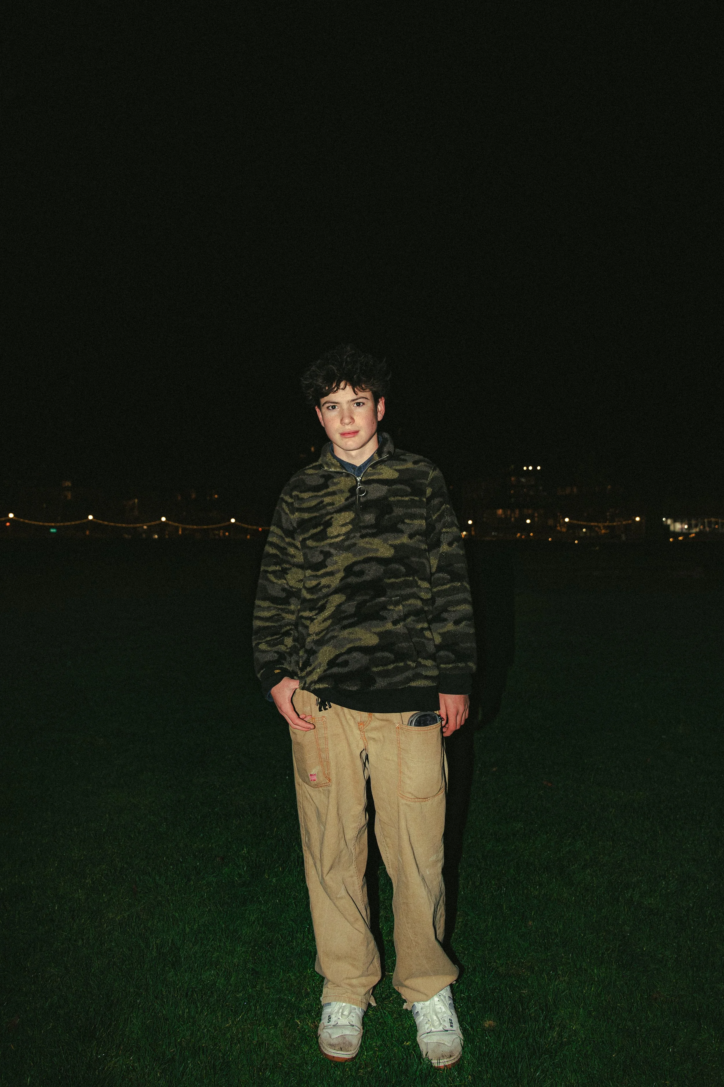 A young person standing on grass at night with city lights in the background, wearing a camouflage jacket, beige pants, and white sneakers.