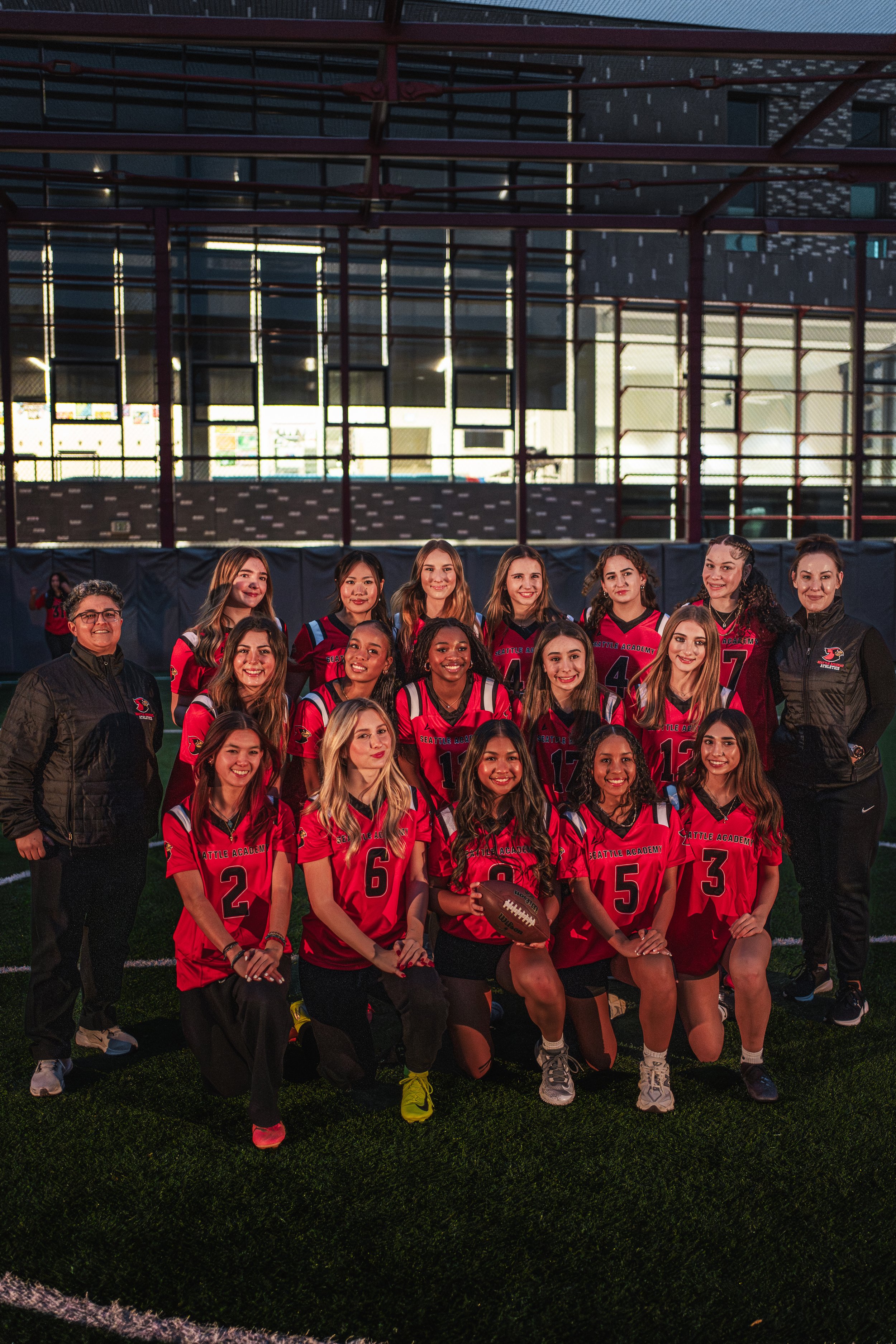 Group photo of a girls' football team in red and black uniforms with coaches, inside a stadium with a large glass window in the background.