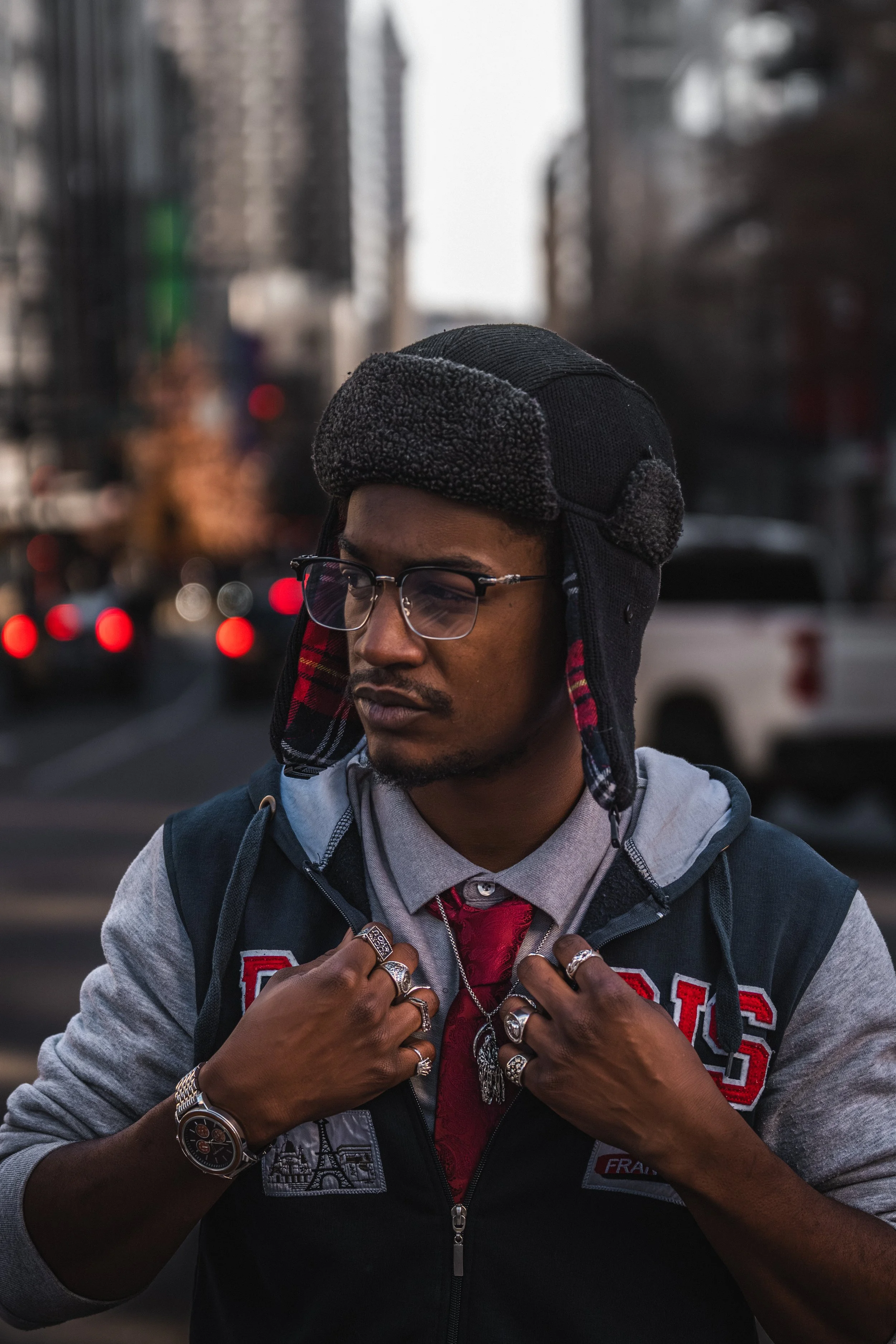 A young man wearing a black and gray winter hat with ear flaps, glasses, a gray hoodie under a dark vest with patches, and multiple rings and a watch, standing outdoors on a city street with blurred cars and buildings in the background.