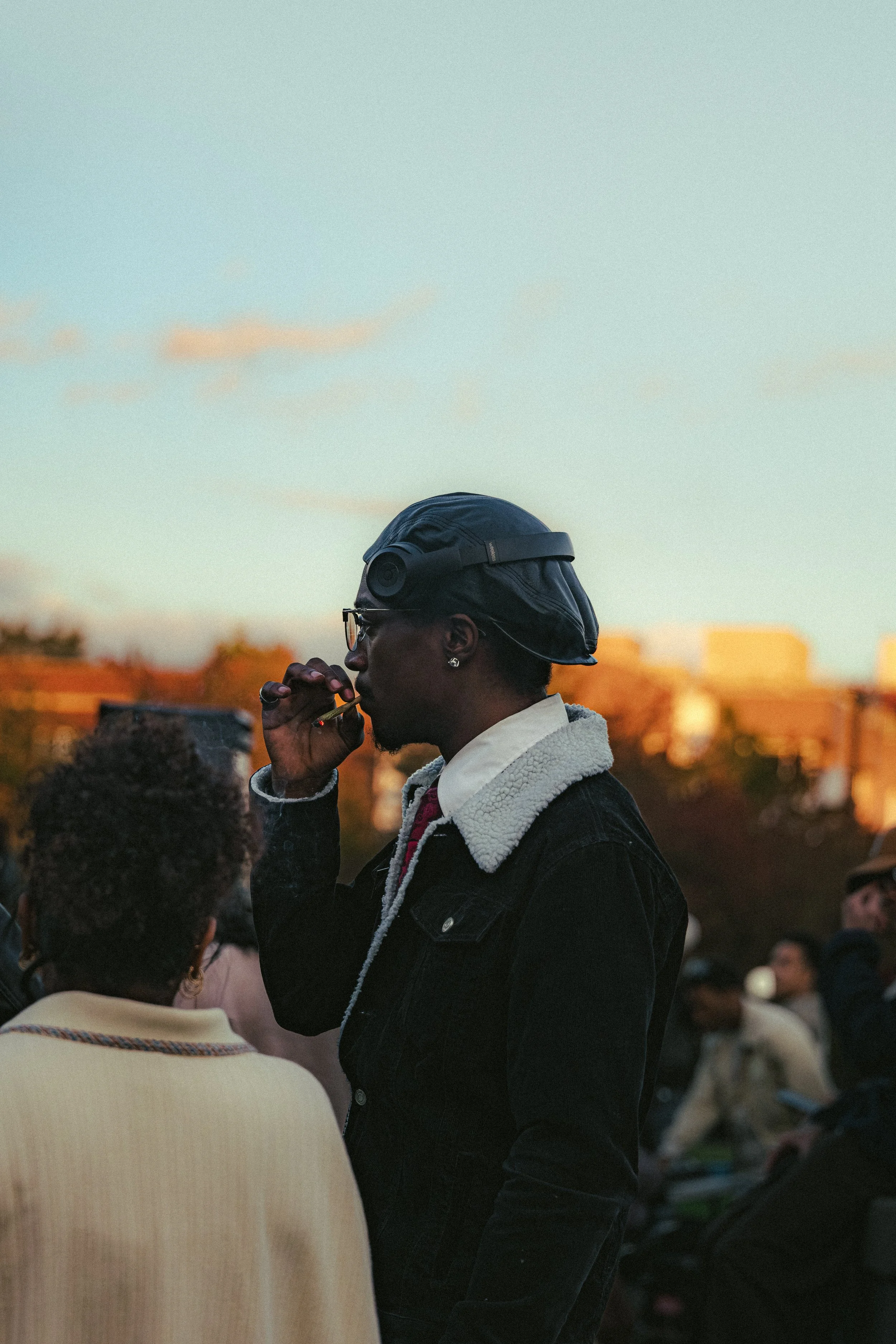 A man standing outdoors, wearing a black cap with a camera attached, smoking a cigarette, with several people in the background during sunset.