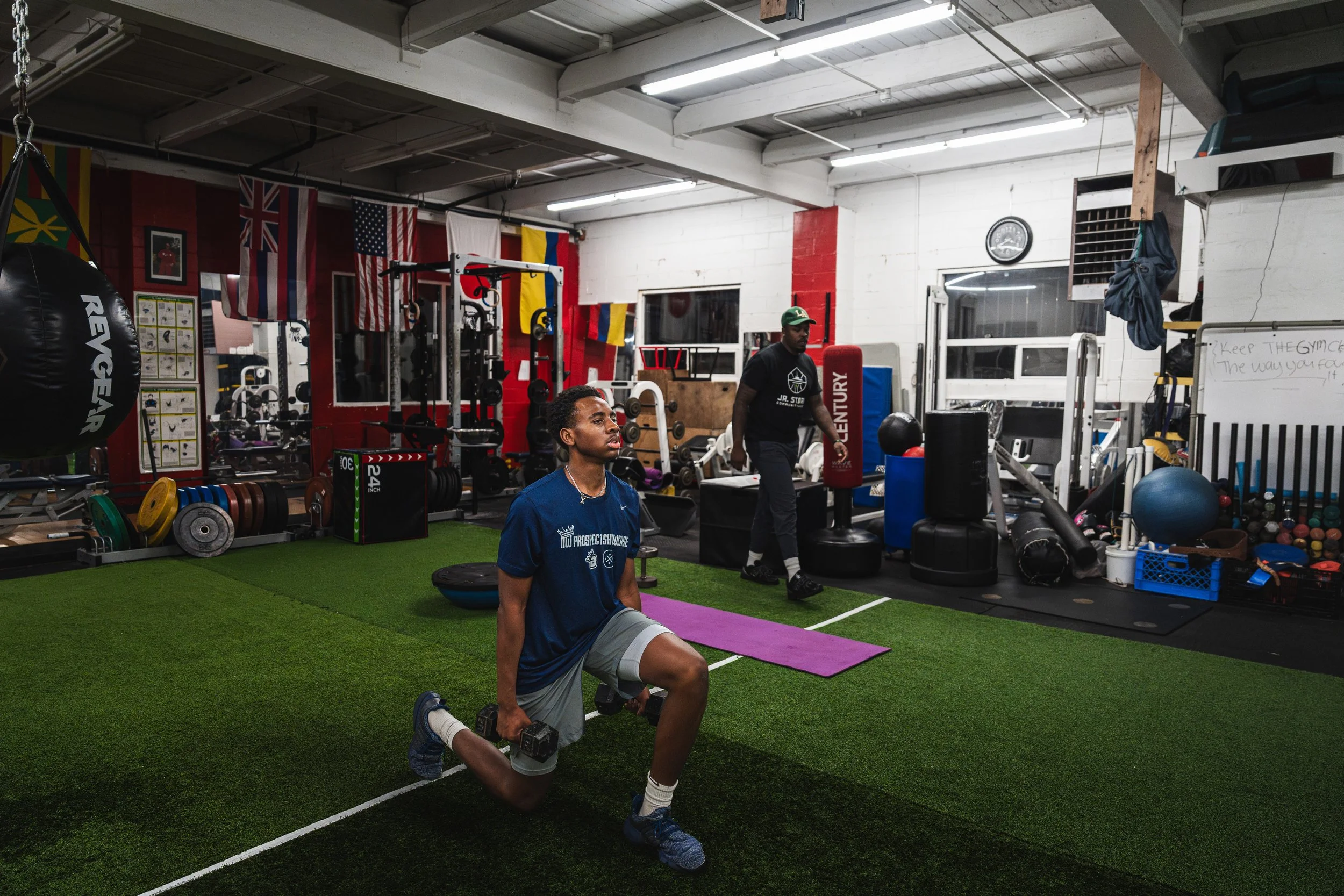 Man performs a lunge with dumbbells in a gym, with weightlifting equipment and flags on the wall in the background, and a trainer standing nearby.