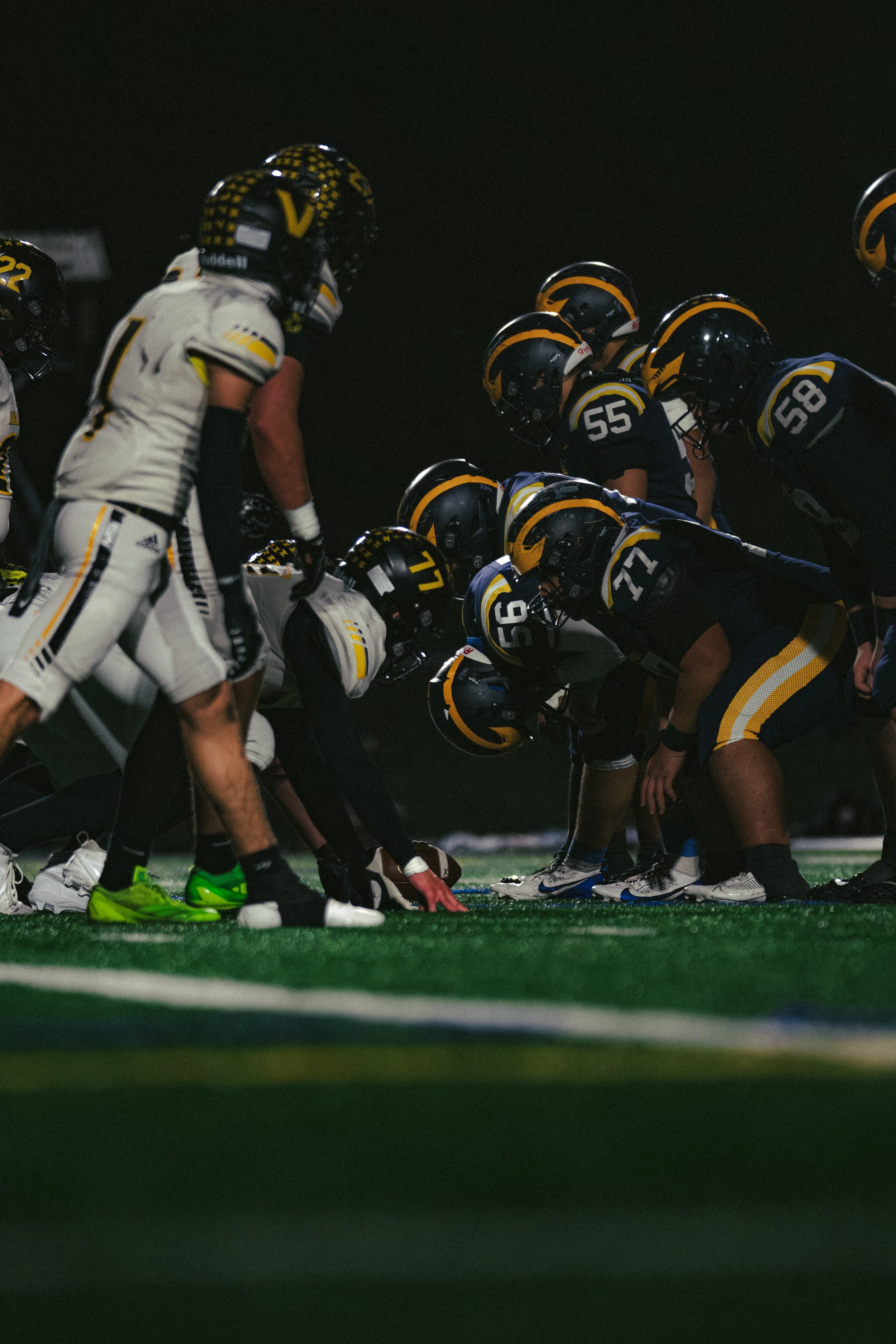 Football players in helmets and uniforms line up at the line of scrimmage at night.