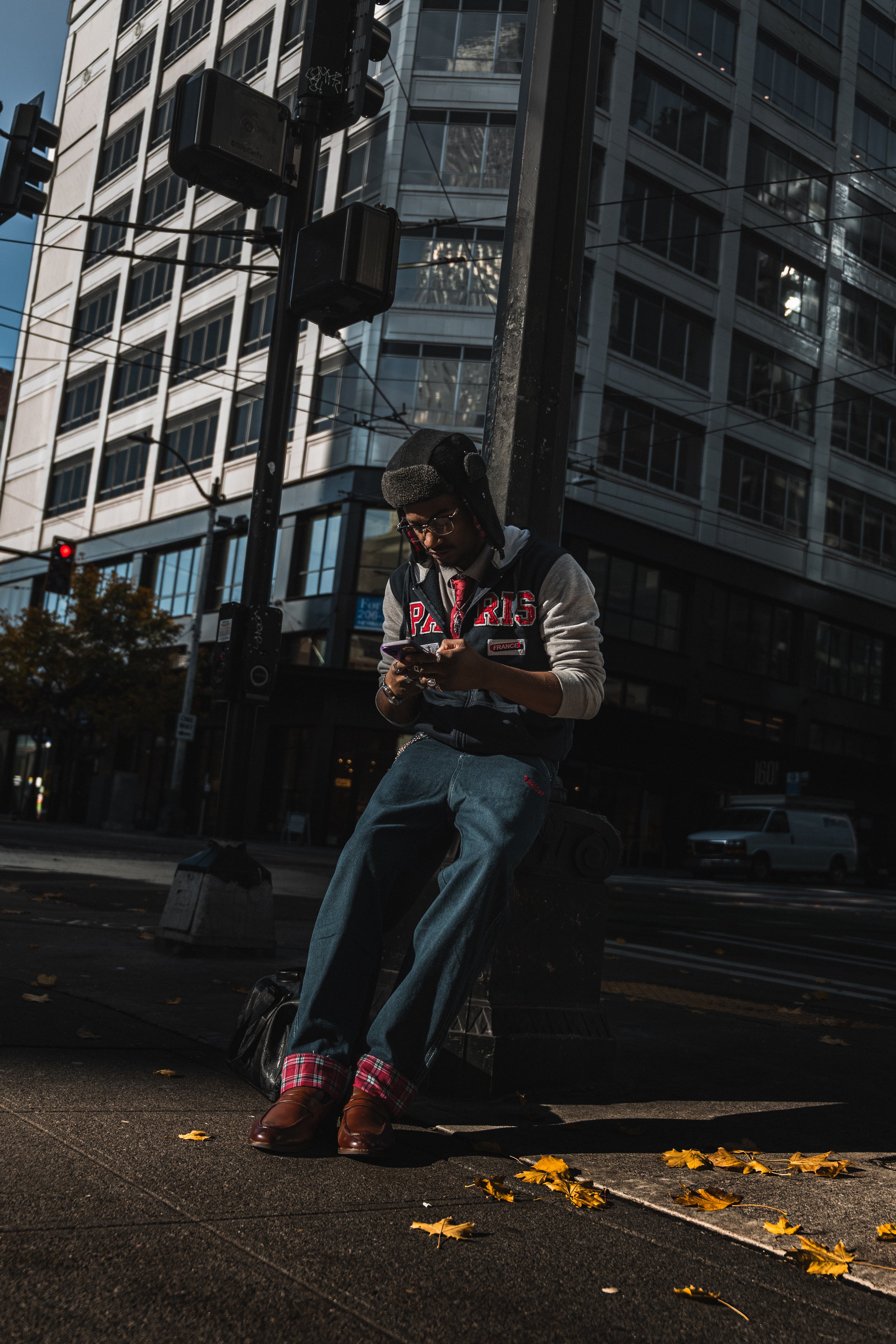 A person sitting on a bench at a city intersection, looking at their phone. They are wearing a gray hoodie with red and black lettering, wide-leg jeans with red plaid cuffs, brown shoes, and a hat with earflaps. The background features tall buildings