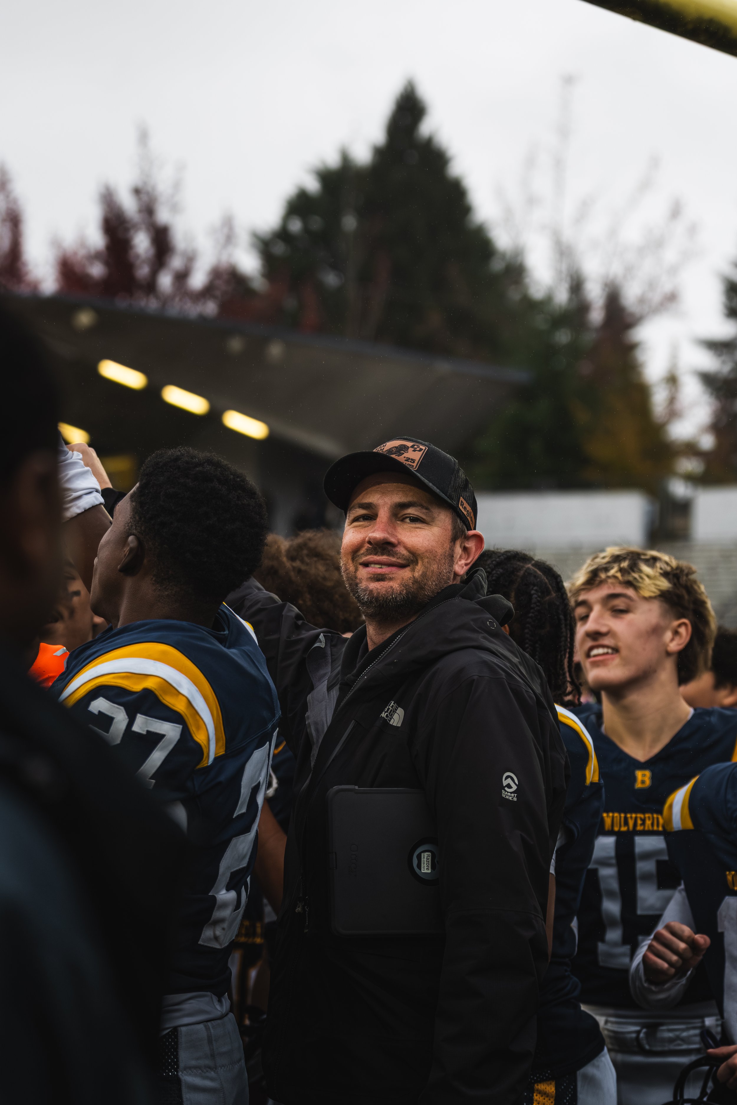 A man wearing a black jacket and cap stands among a group of football players, smiling, with trees in the background.