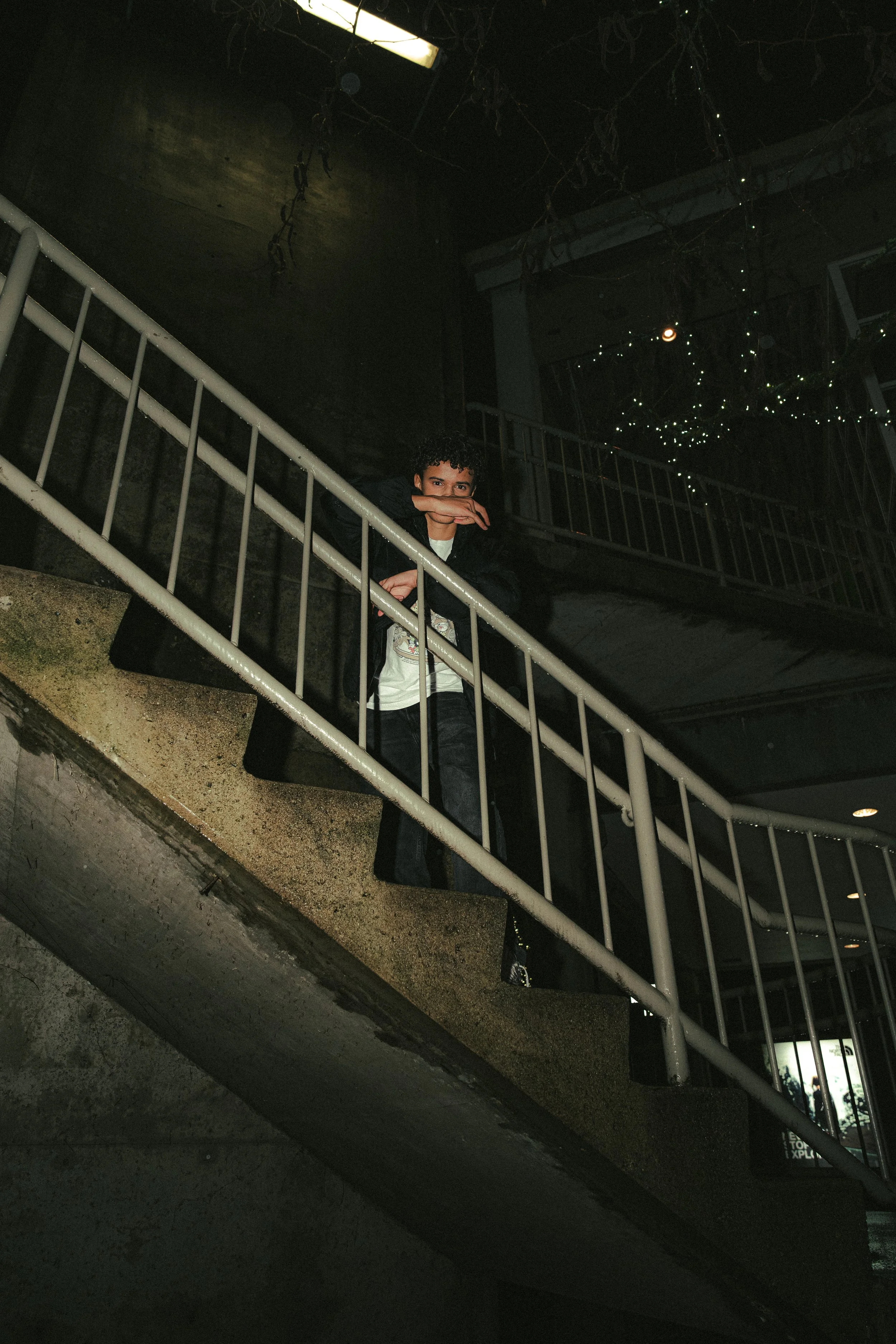 Person standing on a staircase indoors, partially obscured by railing, with dark background and hanging decorative hanging lights.