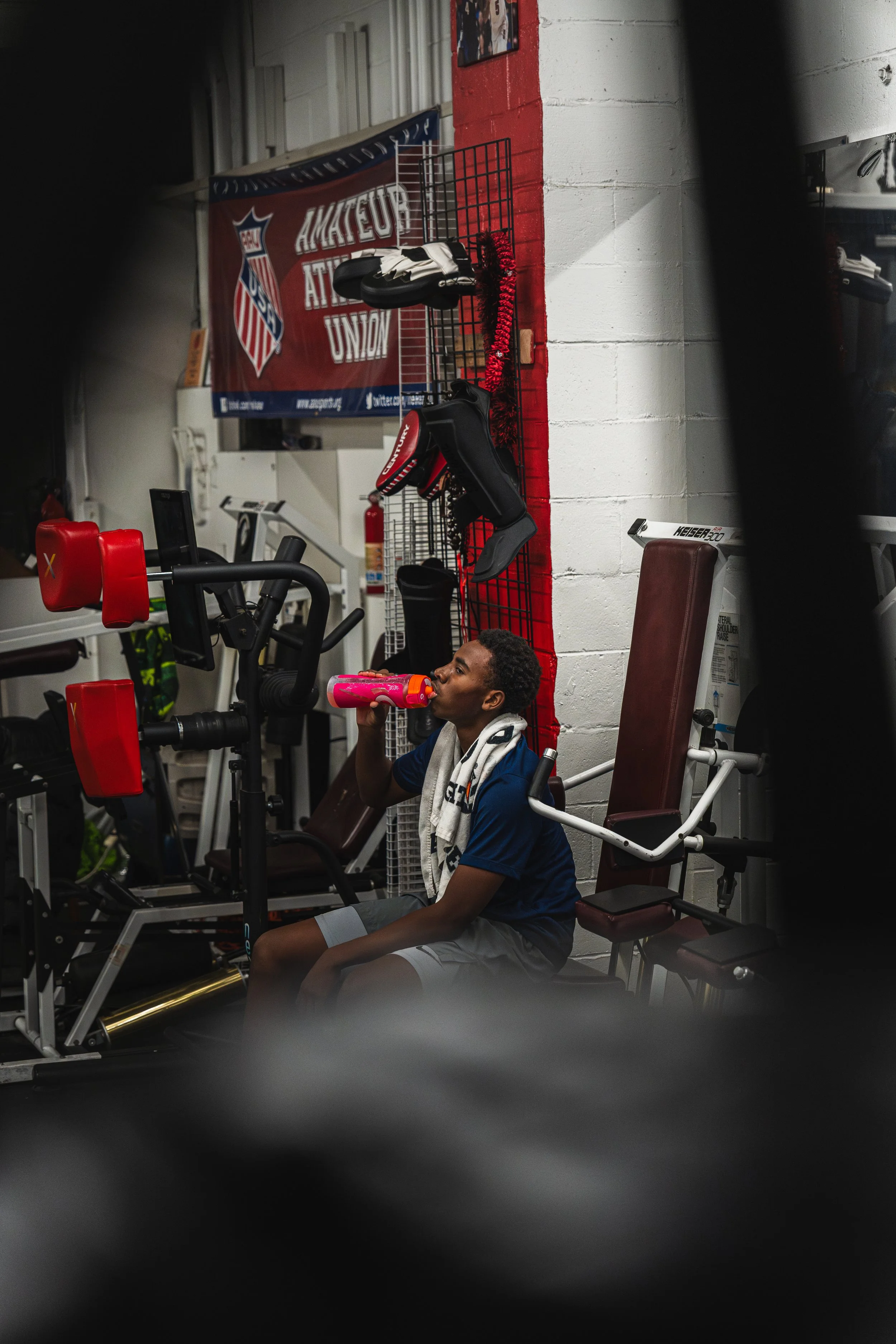 A young man taking a break and drinking from a pink water bottle in a gym, seated on a bench near gym equipment with an 'Amateur Athletic Union' banner on the wall behind him.