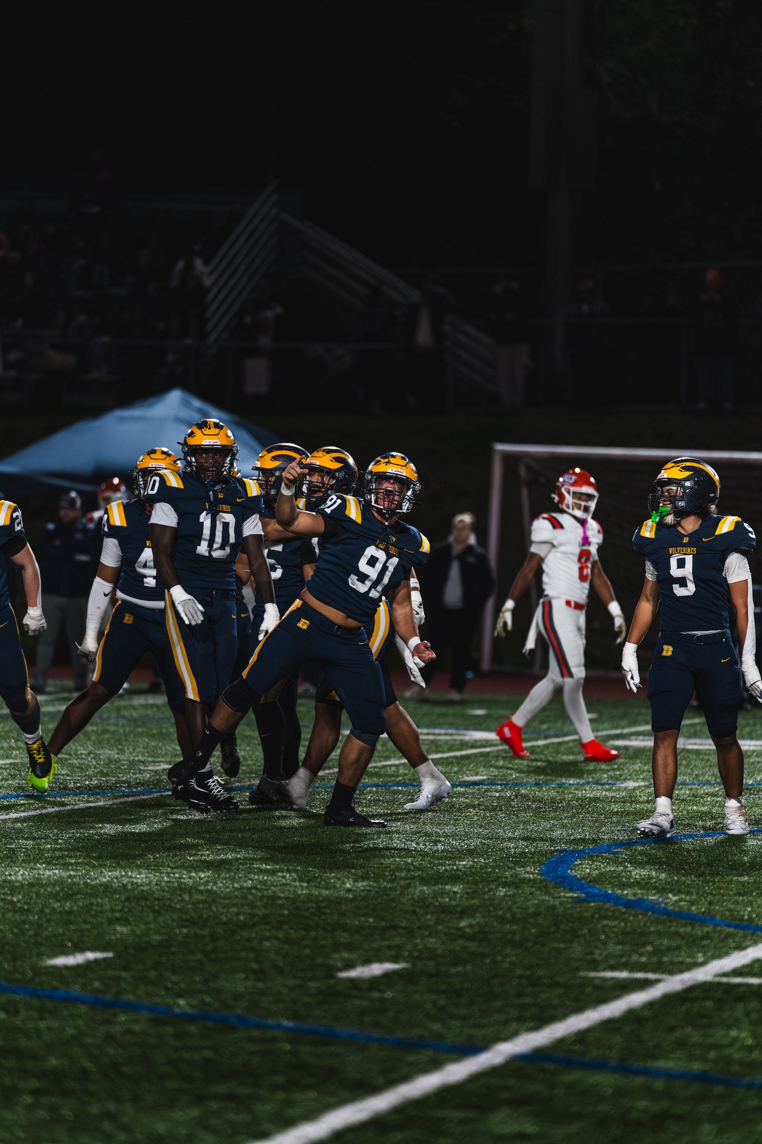 High school football game at night with players in dark blue and yellow uniforms celebrating on the field, and players in red and white uniforms in the background.
