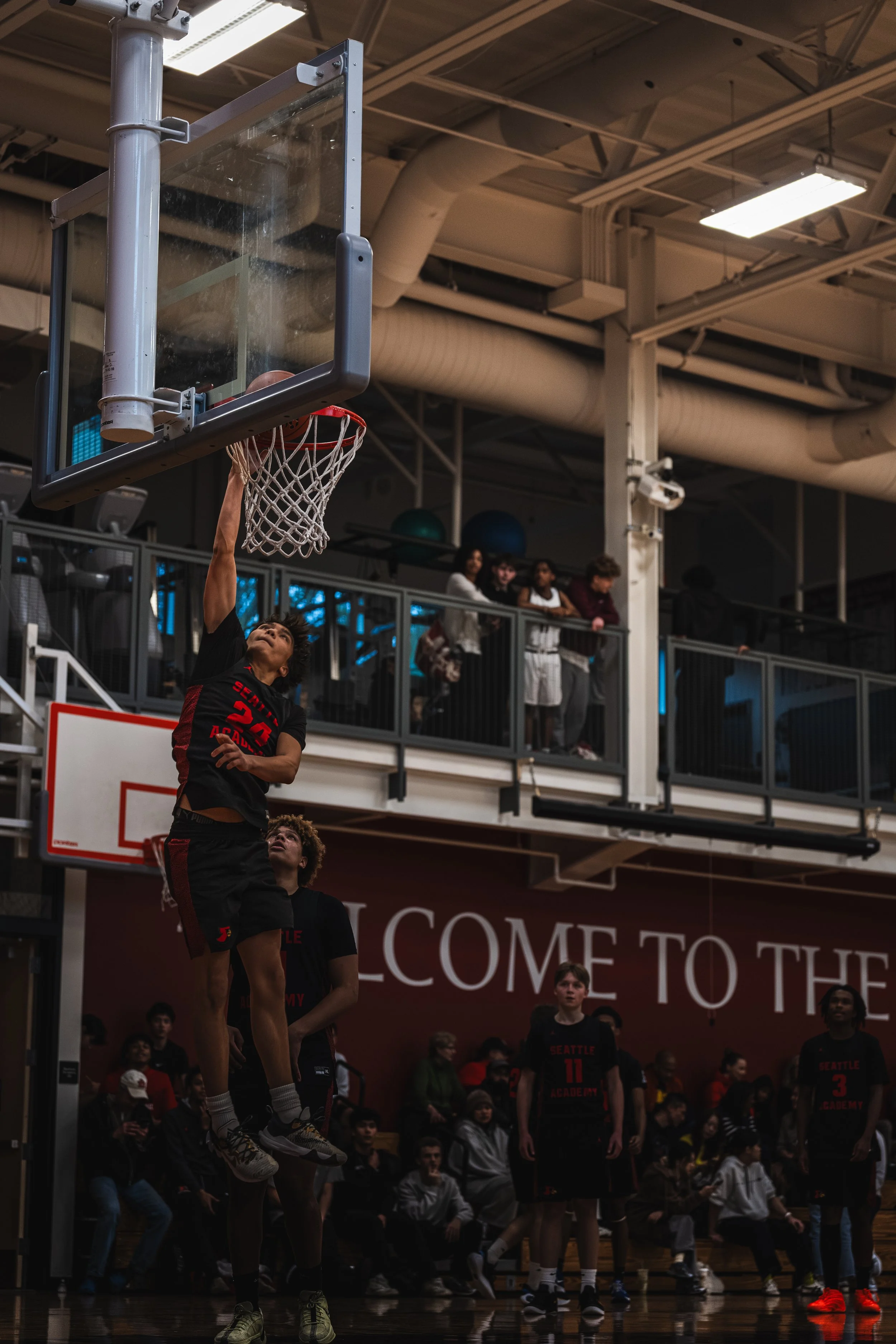 A basketball player in a black and red uniform is jumping to dunk the ball into the hoop, with an audience and teammates watching in the background at an indoor basketball court.