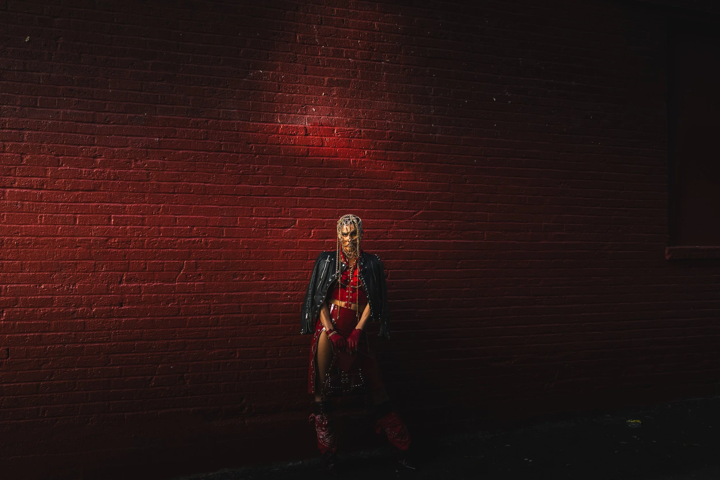 Person with braided hair sitting against a red brick wall at night, dressed in red and black clothing with a leather jacket draped over their shoulders.
