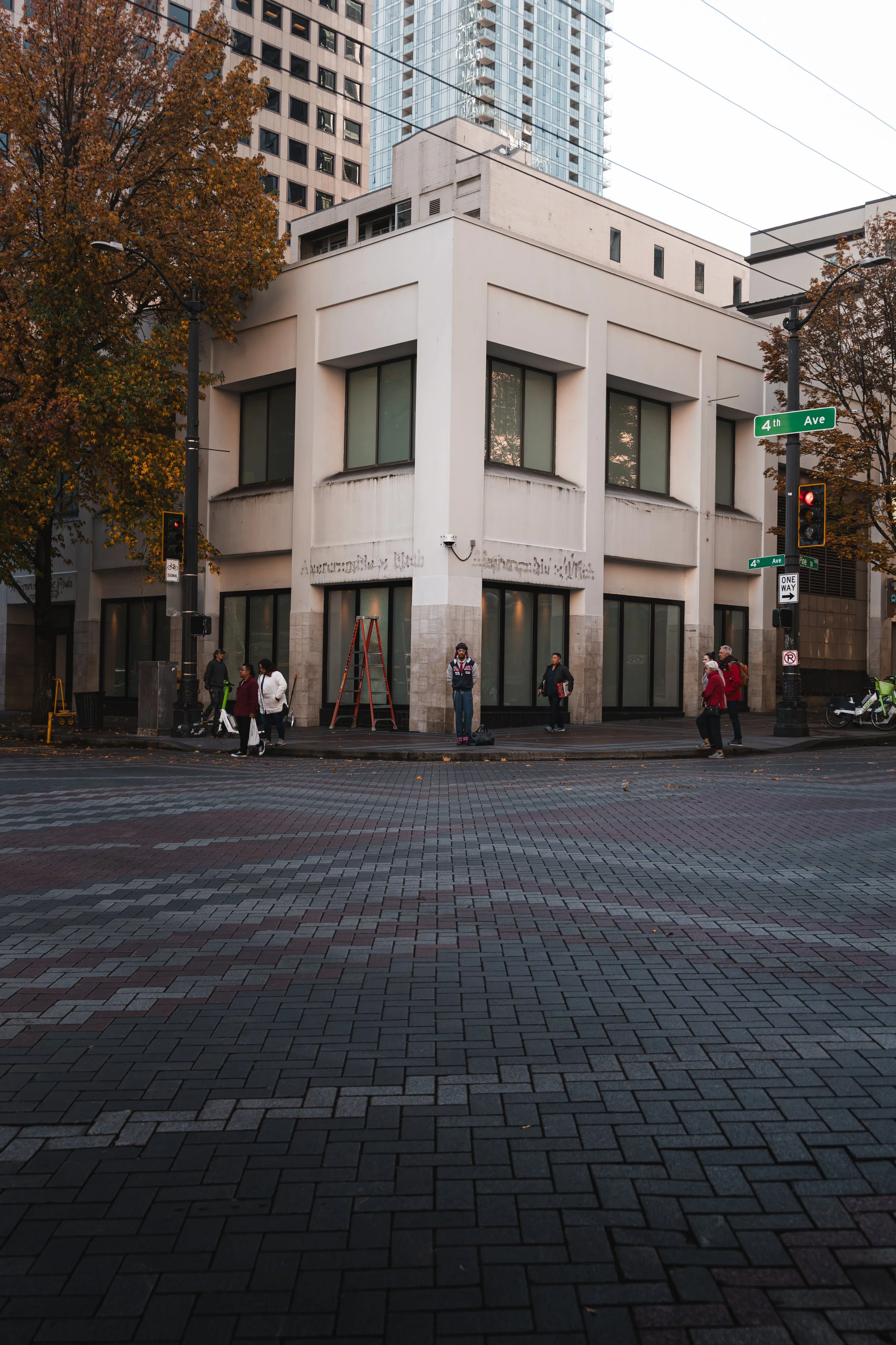 A street corner in an urban area with a white building at the intersection of 4th Ave and an unnamed street. There are trees with autumn leaves and several pedestrians waiting at the crosswalk. A ladder is placed against the building.