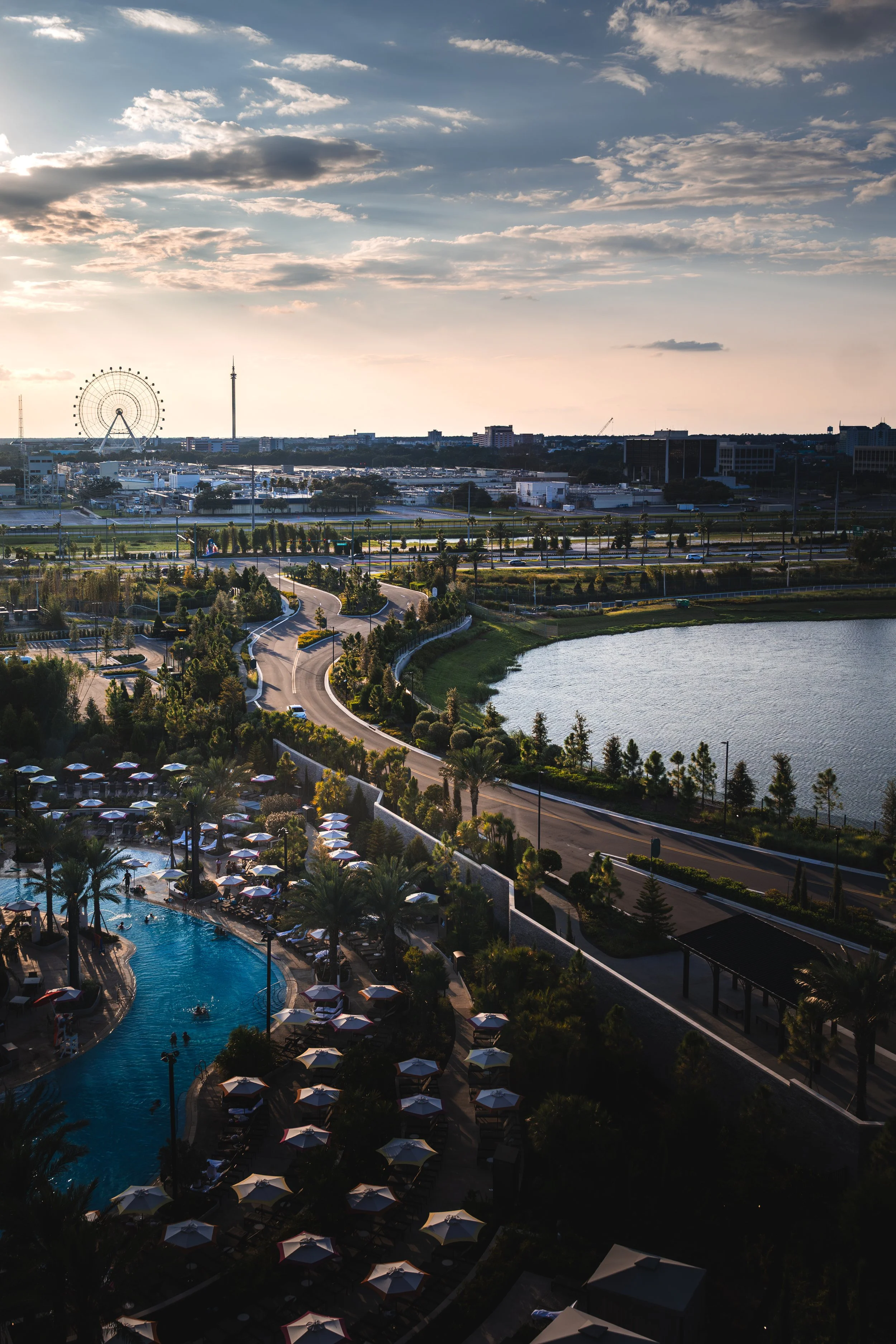 Aerial view of a cityscape featuring a large amusement park ferris wheel, a tower, and a body of water, with a resort pool area surrounded by umbrellas and palm trees in the foreground.