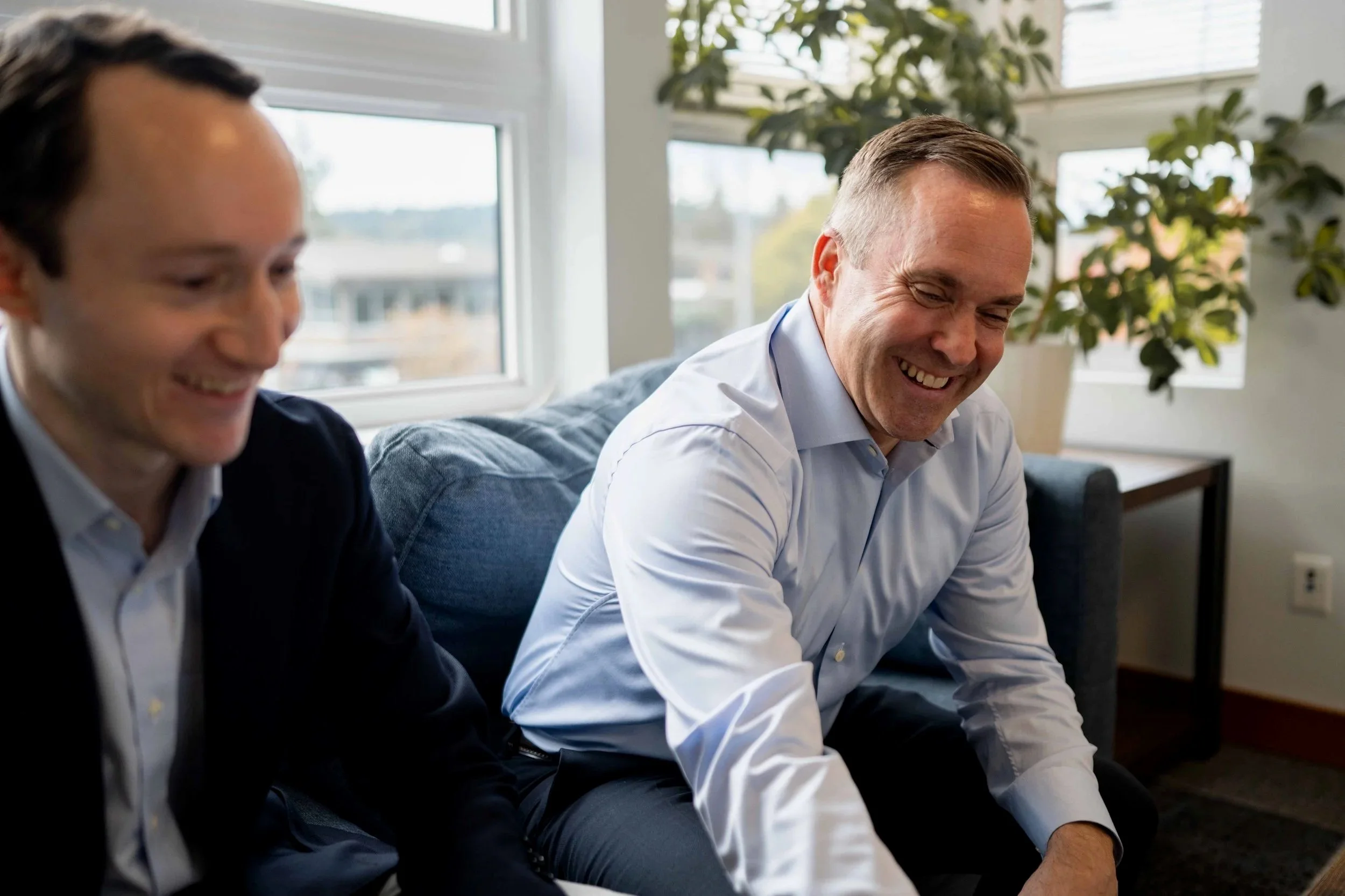 Two men sitting on a couch, smiling and conversing, in a well-lit room with large windows and green plants in the background.