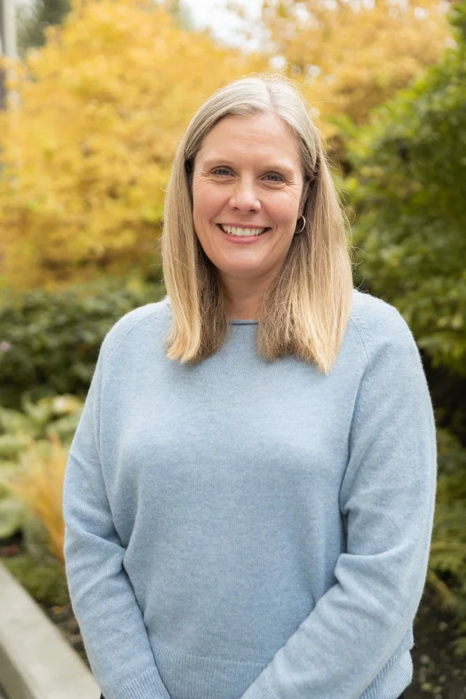 Smiling woman with shoulder-length blonde hair in a light blue sweater standing outdoors in fall foliage.