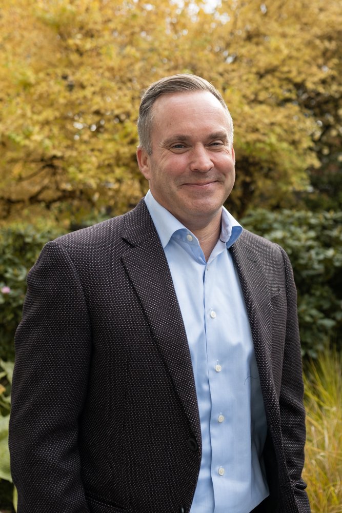 A man wearing a dark, textured blazer and light blue dress shirt standing outside with autumn-colored trees in the background.