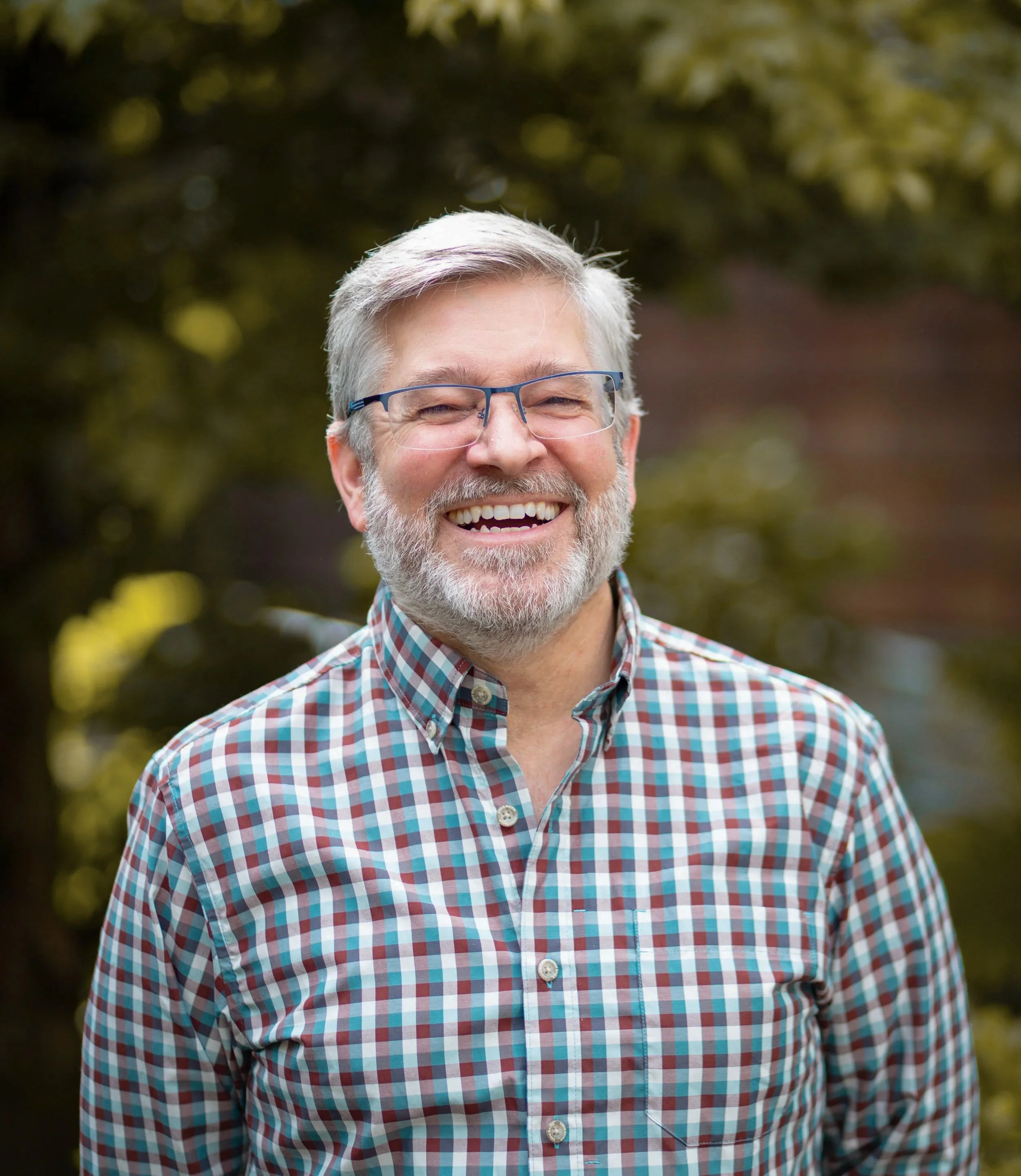 A smiling older man with gray hair and beard, wearing glasses and a colorful checkered shirt, outdoors with blurred trees in the background.