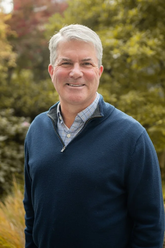 A middle-aged man with gray hair smiling outdoors, wearing a blue quarter-zip sweater over a collared shirt with a blurred green and brown background.